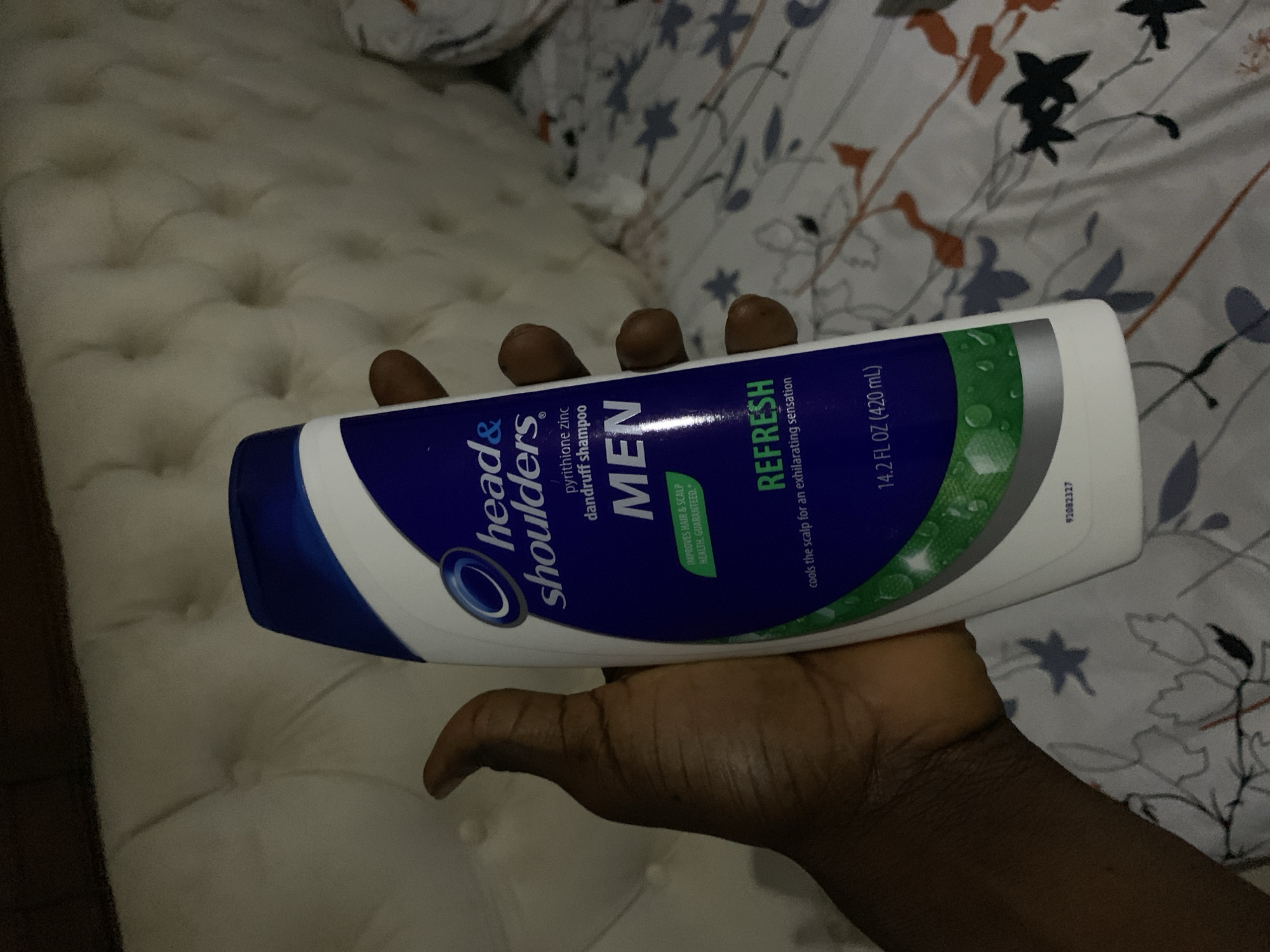 A dark-skinned hand holds a bottle of Head & Shoulders pyrithione zinc dandruff shampoo, specifically the "MEN Refresh" variant, in an indoor setting in Gwarinpa, Nigeria. The white and blue bottle is held horizontally, prominently displaying its label. Visible text on the bottle includes "head& shoulders", "pyrithione zinc dandruff shampoo", "MEN", "IMPROVES HAIR & SCALP HEALTH. GUARANTEED.", "REFRESH", "cools the scalp for an exhilarating sensation", and "14.2 FL OZ (420 mL)". A product code "92082327" is also present.
The background suggests a bedroom environment with dim lighting. A light-colored, tufted surface, resembling a mattress or headboard, is partially visible. Behind this, a patterned fabric, likely a duvet or comforter, features a design of stylized leaves and branches in shades of grey, orange, and blue on a light background. The focus is on the shampoo bottle and the hand holding it, with the background providing context to the scene.