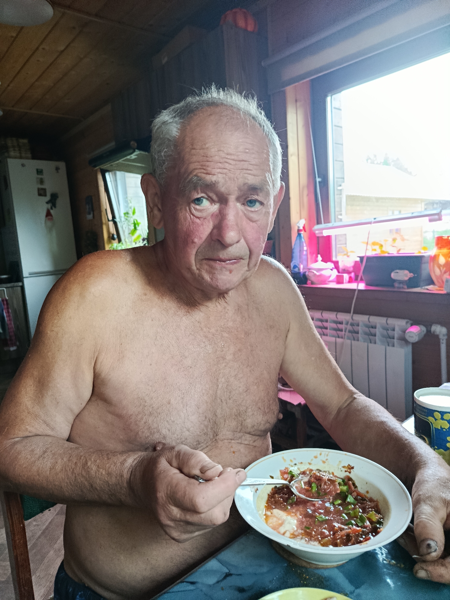 An older man with graying hair and weathered skin sits at a table, shirtless, holding a spoon to a bowl of food. He is looking directly at the camera with a slightly concerned expression. The background appears to be an indoor setting with wooden walls and ceiling, a window with a view of the outside, and various objects on a shelf, including what looks like a refrigerator. The table is covered with a blue tablecloth, and the man is wearing blue shorts. The scene is lit by natural light coming from the window and an artificial light source above.