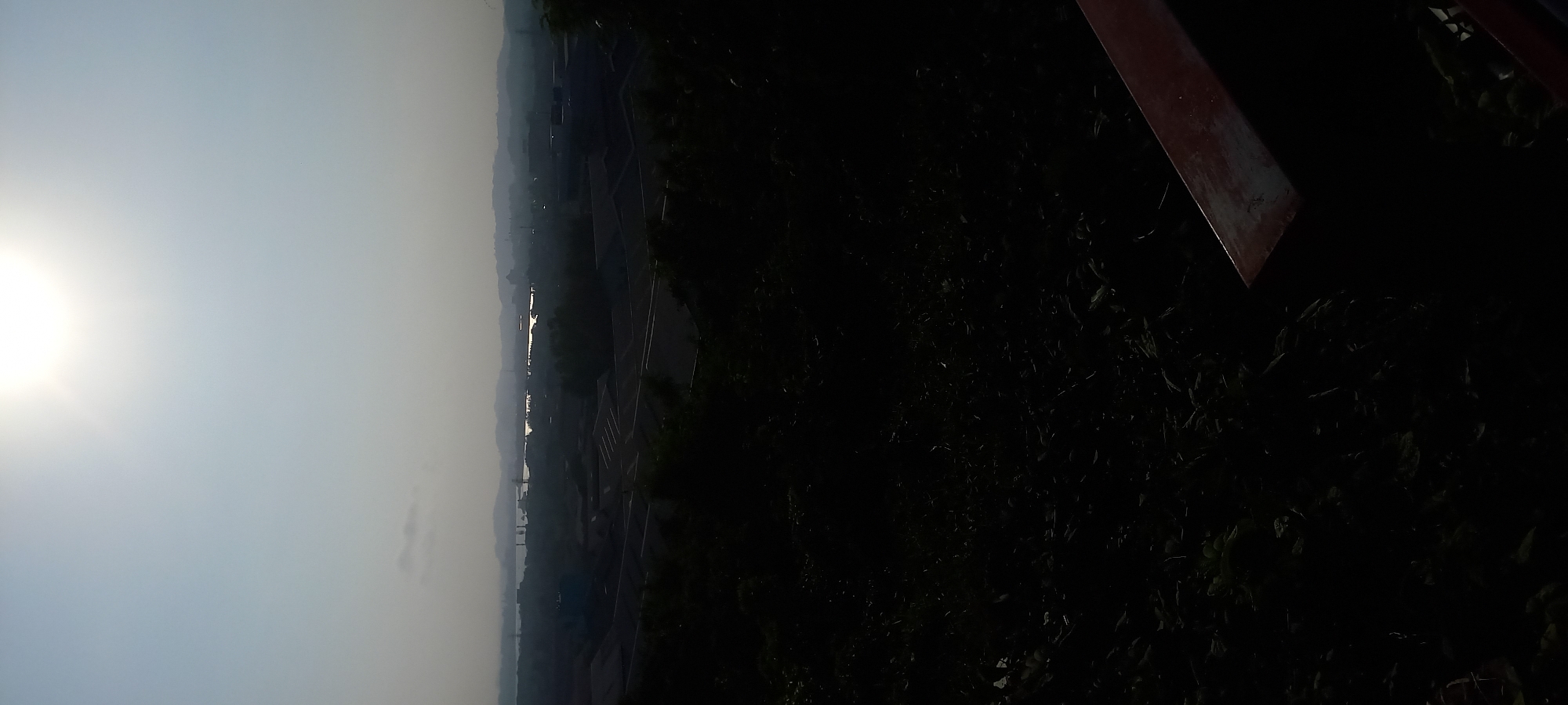 This image captures a panoramic view of Samarinda, Indonesia, from an elevated vantage point. The scene is dominated by a hazy, sunlit sky, with the sun itself appearing as a bright, diffused orb near the top center of the frame. Below the sky, a distant body of water, possibly the Mahakam River, reflects the light and is dotted with structures. Further into the middle ground, industrial-looking buildings with angular roofs are visible, interspersed with darker, silhouetted trees. The foreground is heavily shadowed, obscuring details of foliage and possibly a railing or bench in the lower right corner. The overall impression is one of an industrial or semi-urban landscape under a bright, hazy sky.