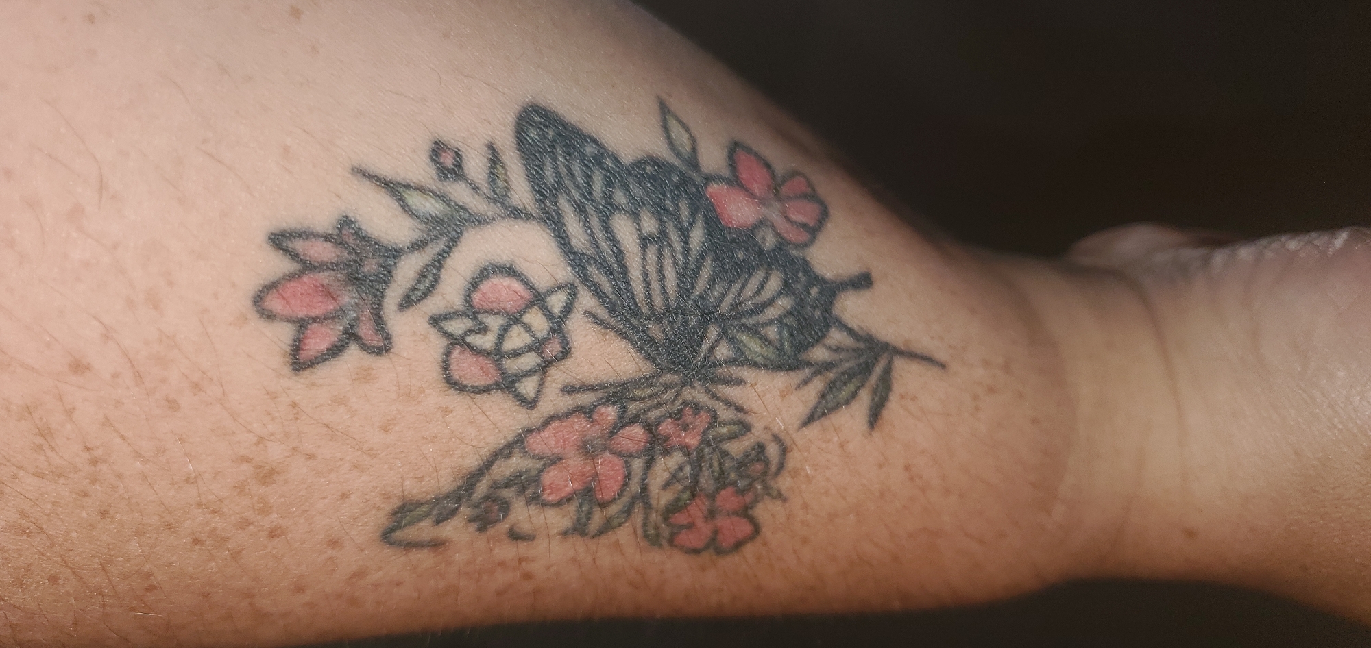The image shows a tattoo on a person's forearm in Sasbach, Germany. The tattoo depicts a butterfly with intricate wing patterns in black ink. Surrounding the butterfly are several small, stylized flowers in shades of pink and red, with green leaves and branches. One of the flowers, located below and to the left of the butterfly, appears to have a geometric pattern within its petals. The skin tone is light with visible freckles. The background is dark and out of focus, suggesting the image was taken indoors with artificial lighting. The focus is entirely on the tattoo, showcasing its details and colors against the skin. There is no visible text in the image. The lighting creates shadows that highlight the texture of the skin and the tattoo itself.