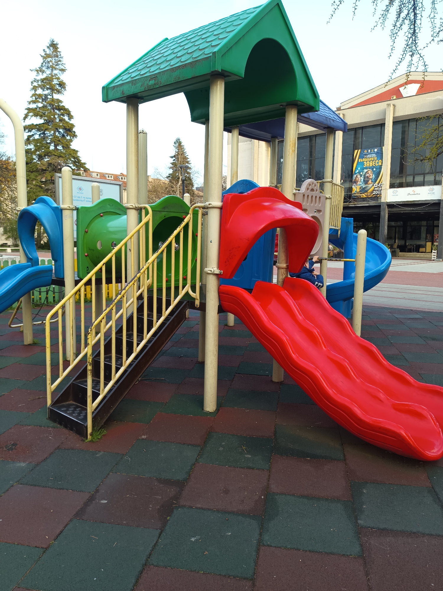 The image shows a playground structure with a green roof, yellow stairs, and a red slide. The structure also features blue and green tunnel elements. The playground is situated on a surface of green and maroon checkerboard tiles. In the background is a building with a banner advertising "Vŭlшебnata Zavesa," visible behind the play structure, which identifies the location as the city of Targovishte, Bulgaria. A small boy is seen inside of the play structure at the top of the slide.