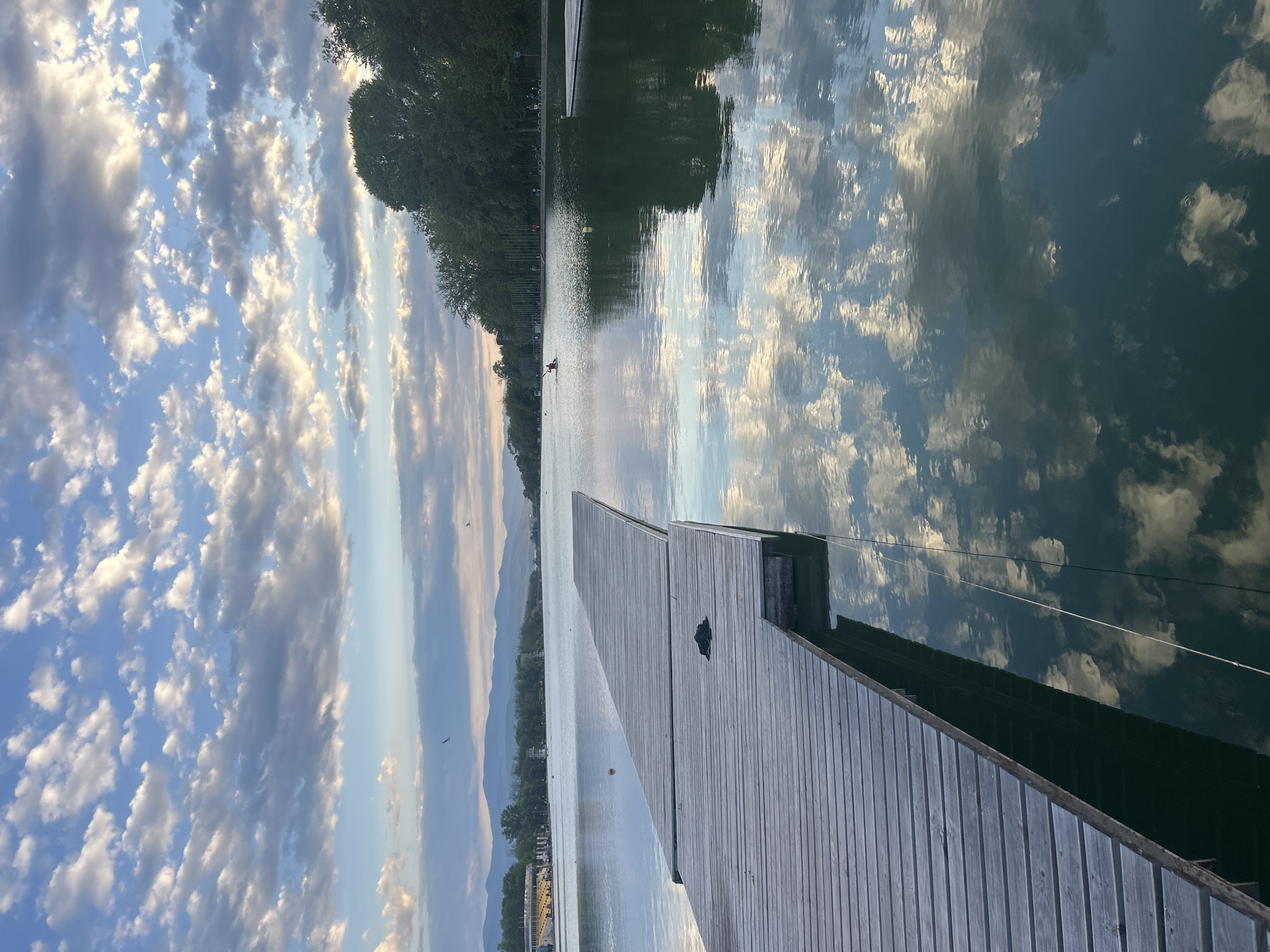 The image depicts a lake scene with a wooden dock extending into the water. The sky features scattered clouds. The water surface reflects the sky and nearby trees. Mountains are visible in the distance. Based on the details in the image and the reference to Plovdiv, Bulgaria, the location is likely the Rowing Canal Plovdiv, a well-known artificial lake used for water sports and recreation. A dark object is visible on the dock. The scene appears calm.