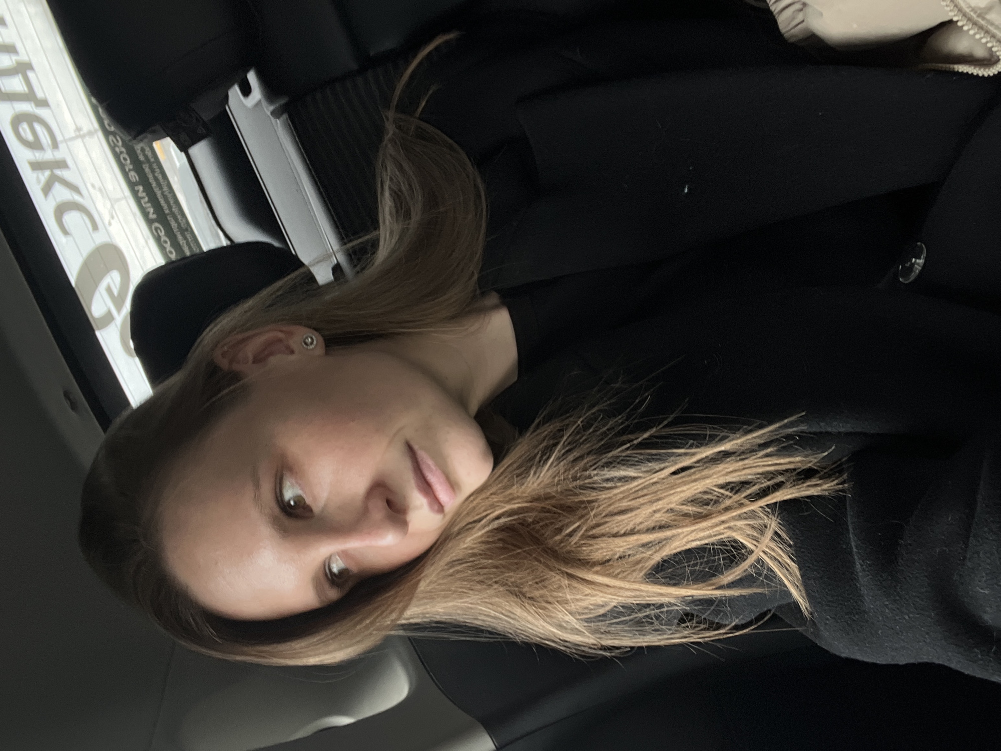 A young woman with light brown hair sits in what appears to be a car. She wears a black sweater. The background reveals a partial view of a window with the text "OKC" printed on it and details of the car's interior. Based on the window's reflection, and the information about location, it can be supposed that the car is in the city of Khimki, Russia.
