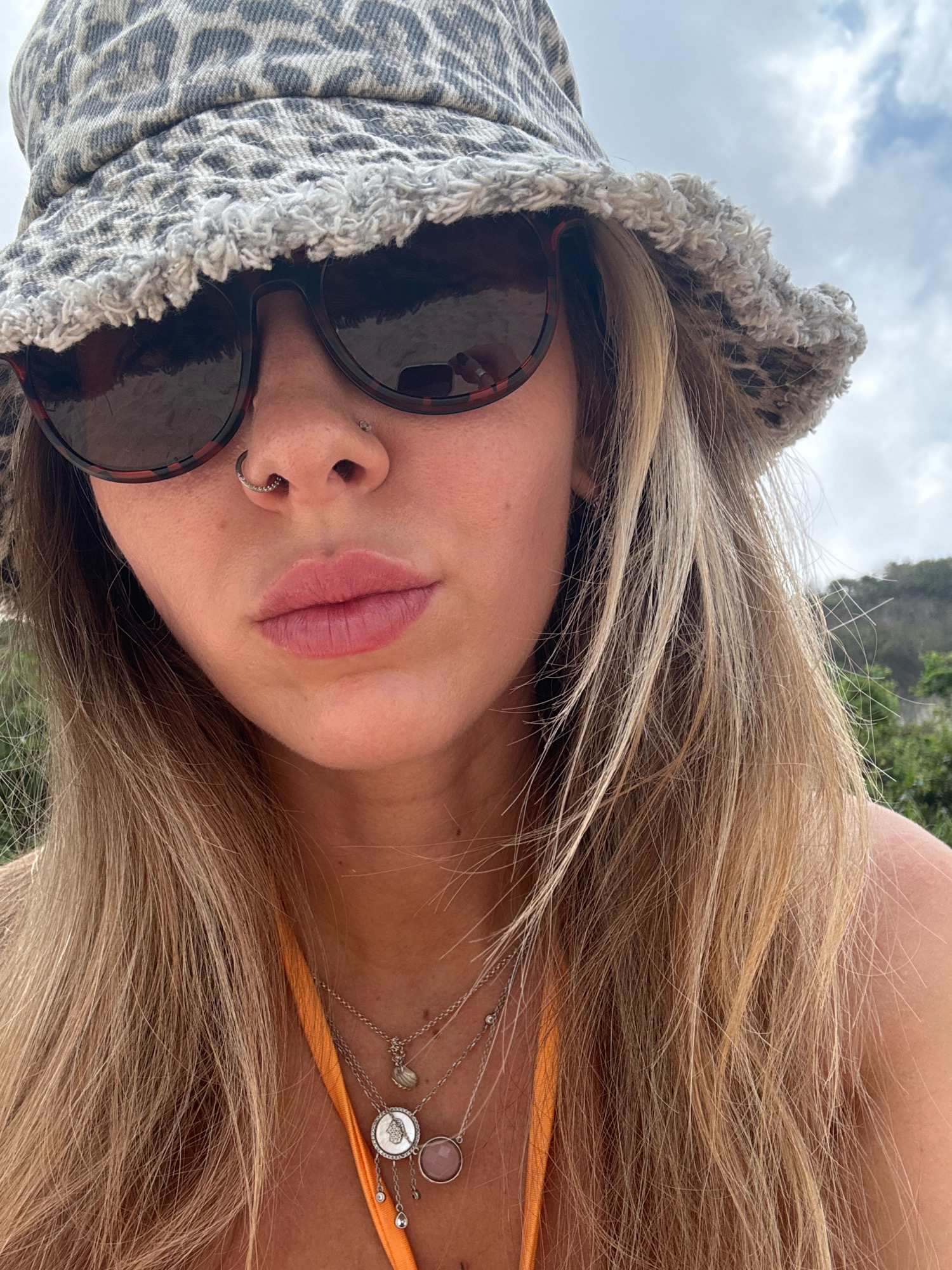 A woman is pictured from the chest up, wearing a leopard-print bucket hat and sunglasses. She has blonde hair and a nose ring. She is also wearing necklaces and an orange bikini top. The background appears to be outdoors, with a cloudy sky and some greenery.