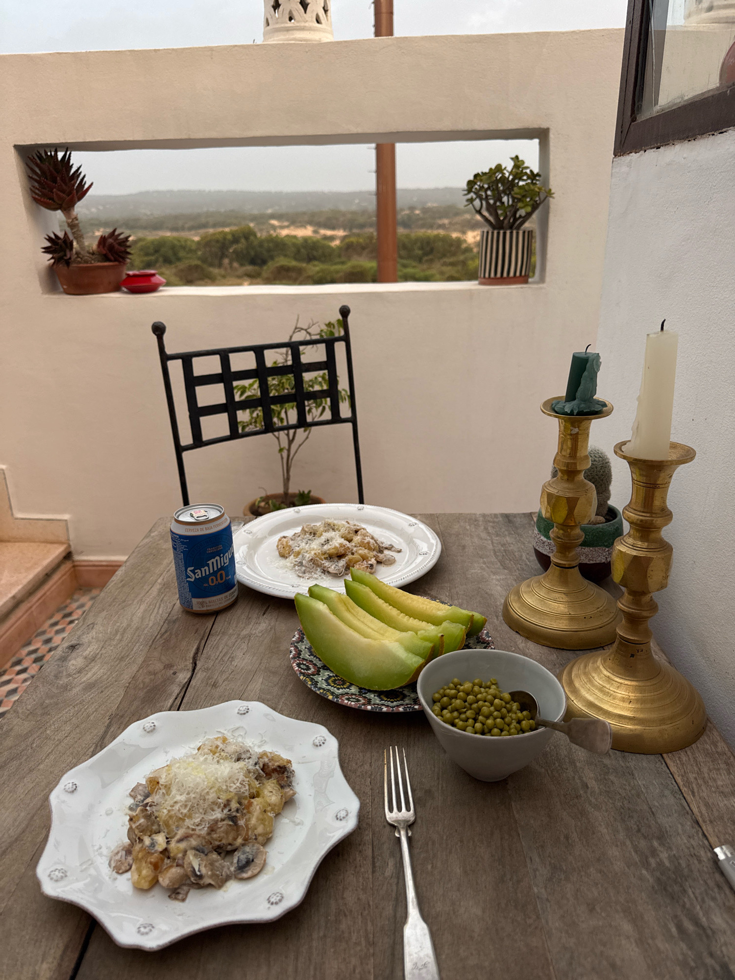 A rustic wooden table is set for a meal on a rooftop patio overlooking a landscape of dunes and vegetation. Two plates of gnocchi with mushrooms and cheese are present. One plate is in the foreground, and the other is further back. Accompanying the food are slices of melon on a patterned plate, a small bowl of green peas with a spoon, and a can of San Miguel beer. A fork rests to the left of the foreground plate. Two brass candle holders with candles, one green and one white, stand on the right side of the table, behind a small cactus. In the background, a black metal chair with a plant behind it is visible. The patio has white walls and a decorative white chimney. The view through a rectangular opening in the wall reveals a hazy landscape. The presence of San Miguel beer and the architectural style suggest the location is likely Essaouira, Morocco.