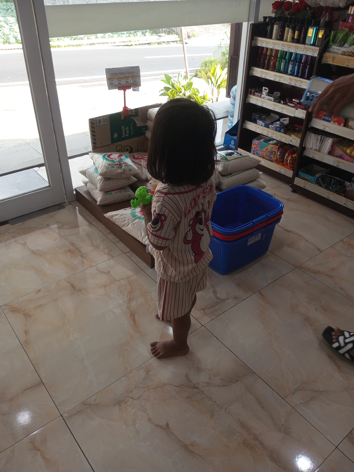 A young child, seen from behind and from the waist down, stands on a tiled floor in what appears to be a small convenience store or market. The child is wearing a striped pajama set with a graphic of a cartoon bear on the back. They are holding a green toy. In front of the child are several sacks of rice stacked on a wooden platform, and a blue plastic shopping basket. To the right, shelves are stocked with various products, including bottles, boxes, and packaged goods. The flooring is light-colored tile with a marbled pattern. Through a large window or glass door in the background, greenery and a street are visible. A price tag is displayed above the rice sacks.