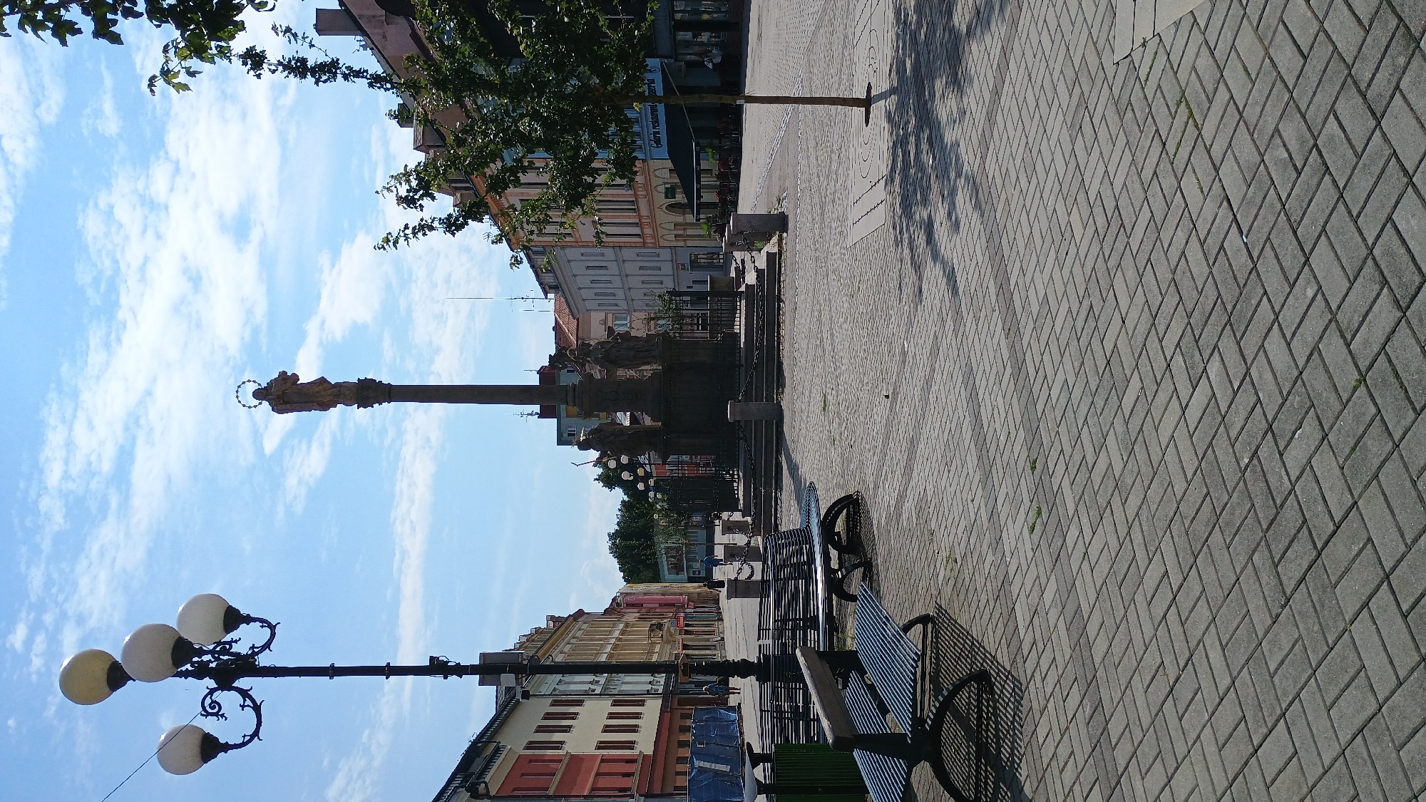 The image shows a street scene in Sokolov, Czechia. The photo is taken in a vertical orientation, with the sky occupying the upper portion of the frame and the street and buildings below.
The scene features a paved plaza with a lamppost and a monument. The lamppost has multiple globes and ornate metalwork. The monument appears to be a column topped with a statue of a figure wearing a halo. The base of the monument is surrounded by a low metal fence, and there are other statues or sculptural elements at its base. Benches are placed along the sidewalk, with one bench in the foreground showing a distinctive metal design and casting a shadow from a tree.
Buildings with colorful facades line the street, suggesting a town square or commercial area. The architecture appears to be historic. There are some trees with green foliage visible, some casting shadows on the pavement. The sky is blue with scattered white clouds.
The image appears to be taken on a sunny day, likely late morning or early afternoon, given the shadows. There is no clear indication of people or specific activities, giving the impression of a quiet moment in the urban environment. The overall mood is peaceful and serene. No visible text is legible in the image.