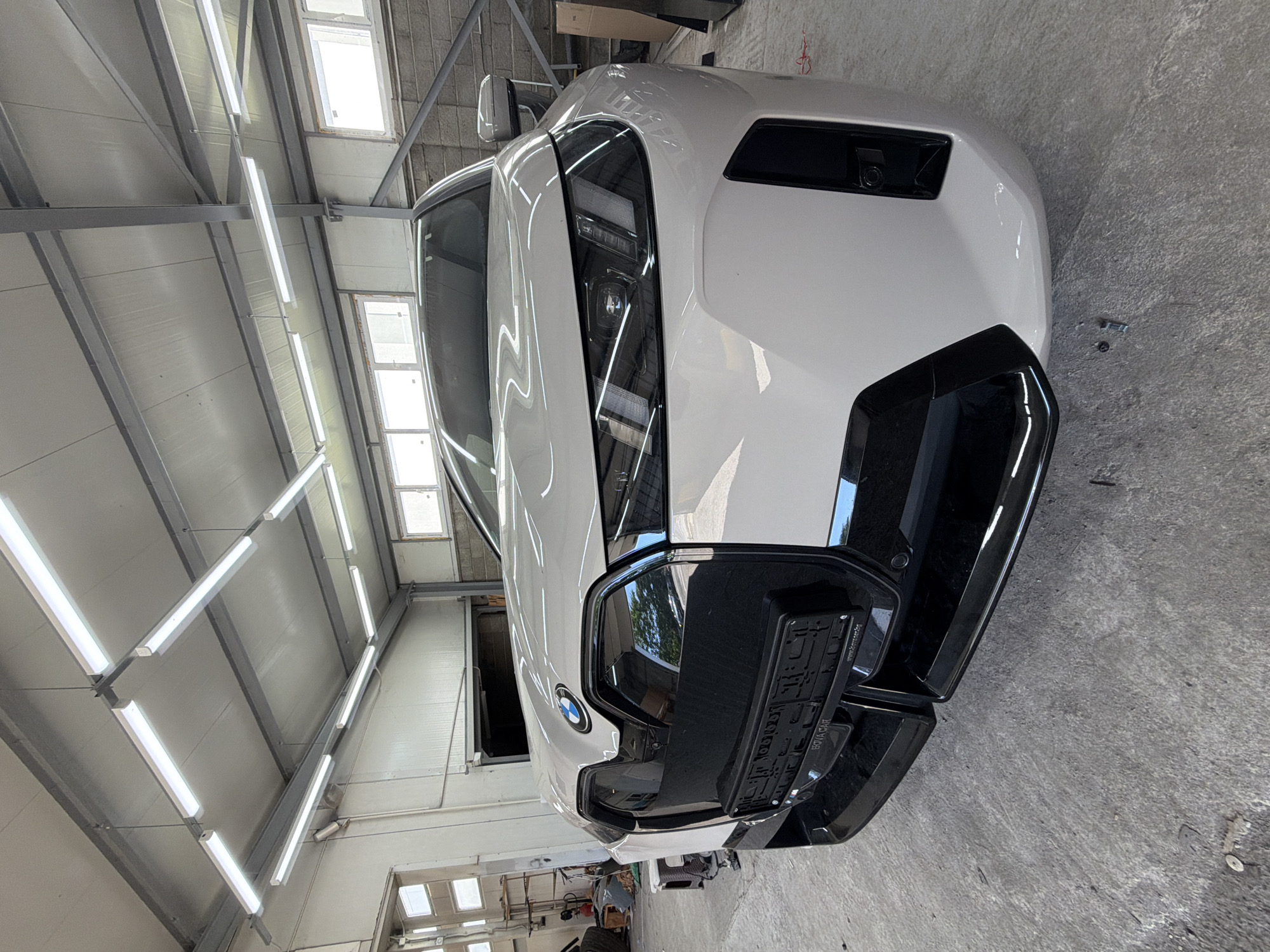 A white BMW iX electric vehicle is parked in a garage with a concrete floor and a metal ceiling with fluorescent lighting. The car is facing the camera, and its front grille, headlights, and license plate are visible. The garage appears to be a workshop or service center, with tools and equipment in the background. The license plate frame has text that reads "BOVA CAR" and "www.bovacar.bg", indicating the location of a car dealership or service in Burgas, Bulgaria.