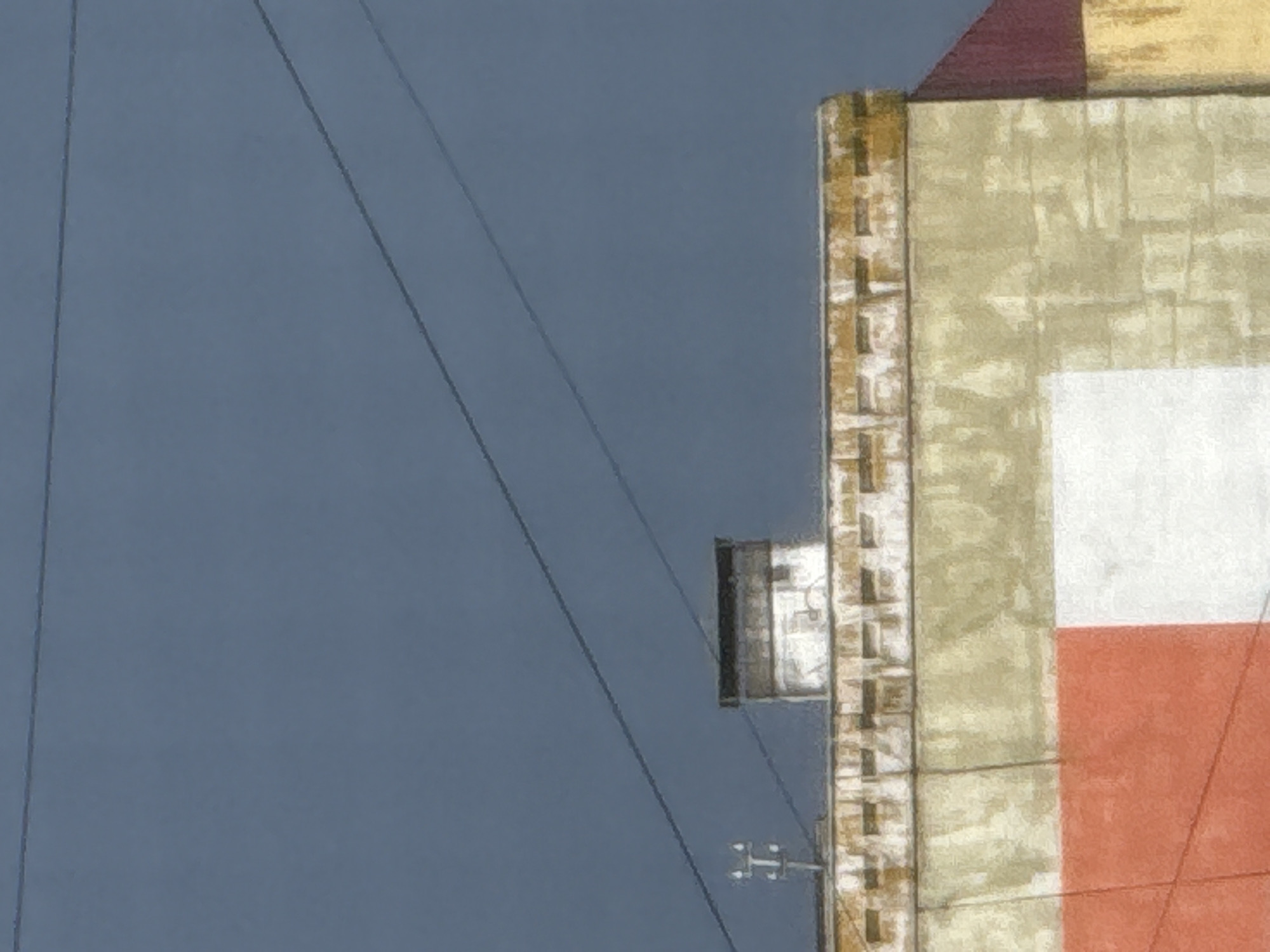 The image shows a close-up of the upper facade of a building against a dark blue sky with thin, dark power lines stretched across it. The building's exterior has a textured appearance, with what looks like brickwork along the top edge, painted in muted yellow and white. Below this, the wall is primarily a pale yellow, with a large section of red on the lower left, and a white section next to it. A red roofline is visible on the right side of the frame. A white chimney with a dark top stands on the roof. On the left side, a small, metallic structure with a "T" shape is visible above the building's facade, likely a utility pole. The scene appears to be in an urban or semi-urban environment in Морозовка, Russia, given the presence of utility infrastructure and building architecture. There are no people visible in the image.