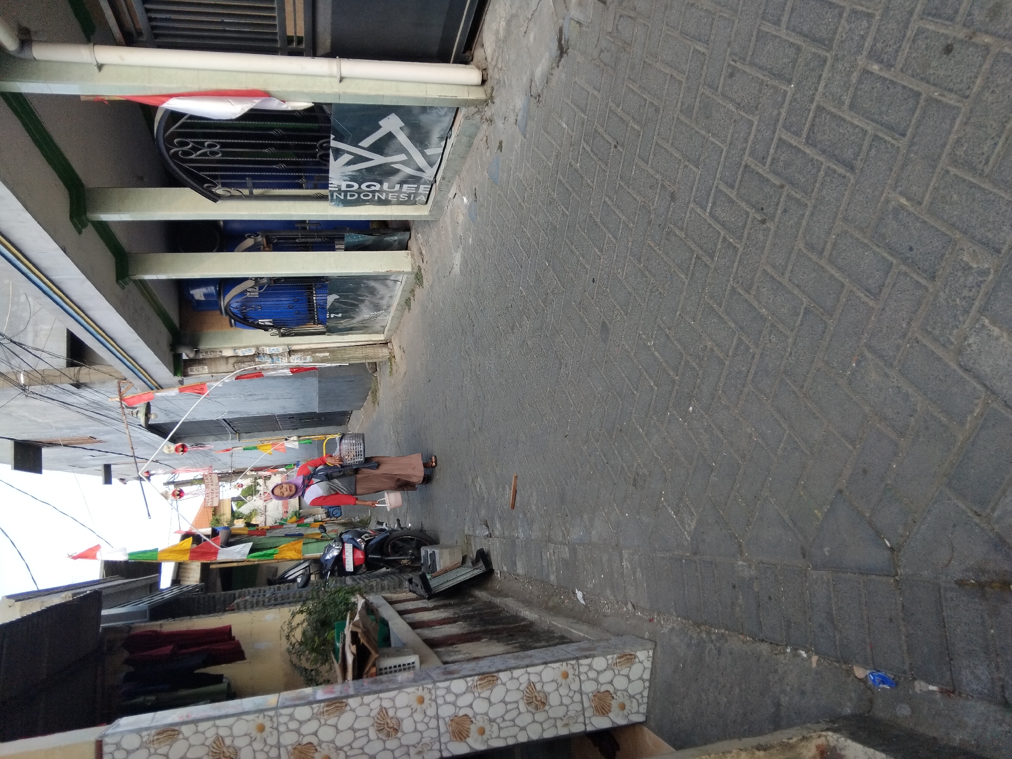A woman wearing a pink headscarf and an orange and grey long-sleeved shirt, with a long brown skirt, walks down a paved alleyway in Kepuh Kiriman, Indonesia. She is carrying a basket in her right hand and a white bucket in her left hand. The alleyway is lined with buildings on either side, with some of the buildings decorated with colorful flags. There is a motorcycle parked on the left side of the alley, and laundry is hanging on a clothesline above it. The ground is paved with dark grey interlocking bricks. In the background, there are large blue containers visible behind a black wrought-iron gate. The scene appears to be during the daytime, with natural light illuminating the area.