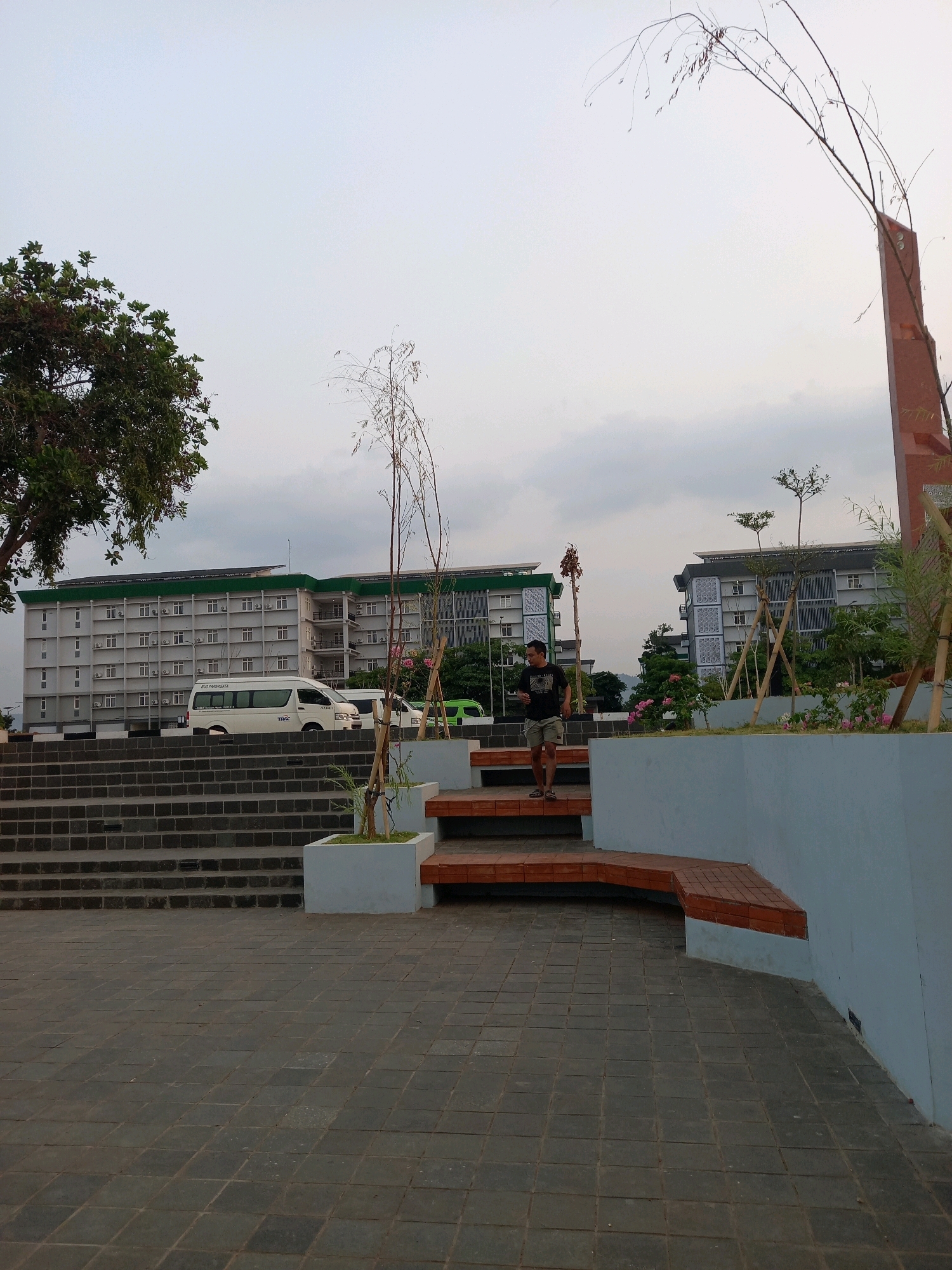 A man wearing a dark t-shirt and shorts walks down a set of stairs in a park area. The stairs are made of dark grey stone blocks. To the right of the stairs, there is a light blue wall with a bench made of reddish-brown tiles. Beyond the stairs and wall, two multi-story buildings with white facades and green roofs are visible. A white van is parked in front of one of the buildings. The sky is overcast. The scene is set in the city of Ketanggan, Indonesia.