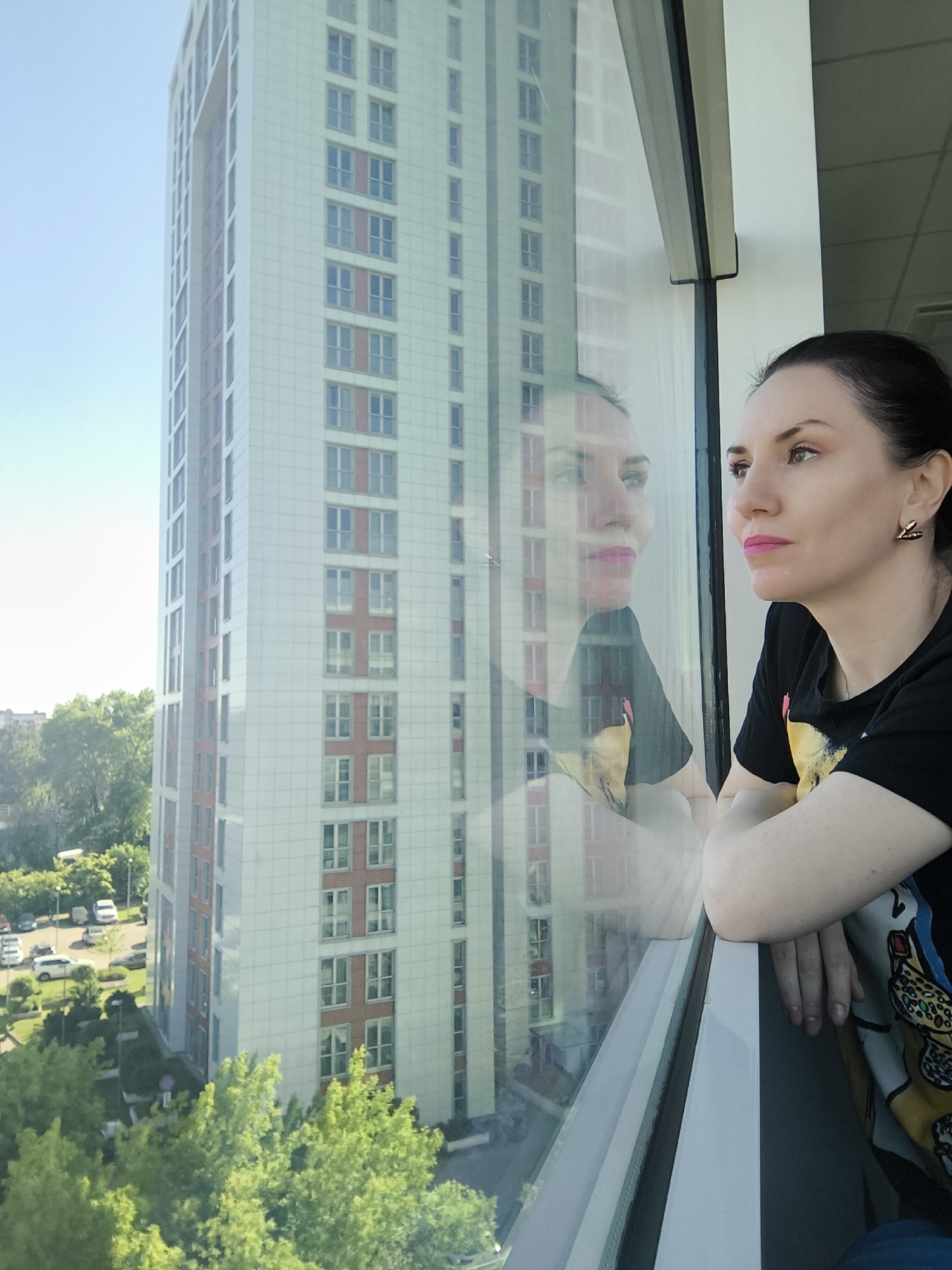 A woman with dark hair pulled back and wearing a black t-shirt with a colorful print is looking out of a large window. Her face is in profile, gazing towards the right side of the frame with a contemplative or observant expression. She has light skin, visible makeup including pink lipstick, and is wearing a geometric gold-toned earring on her right ear. Her forearms are resting on the white frame at the bottom of the window.
A clear reflection of her face and upper body is visible in the window glass, superimposed over the outdoor scene.
Outside the window, a tall, modern building with a light-colored (likely white or light grey) facade dominates the view. The building features numerous rectangular windows, some of which are accented with reddish-brown panels. To the left of this building, another section of a similar structure is partially visible.
Below the buildings and closer to the foreground, a vibrant green canopy of trees and other greenery can be seen. Further in the distance, partially obscured by the trees, there is a parking lot with several cars.
The scene appears to be set during daytime, with clear, bright skies suggesting sunny weather. The vantage point is from a high floor in a building, looking down and across at an urban or suburban landscape. There is no visible text in the image.
