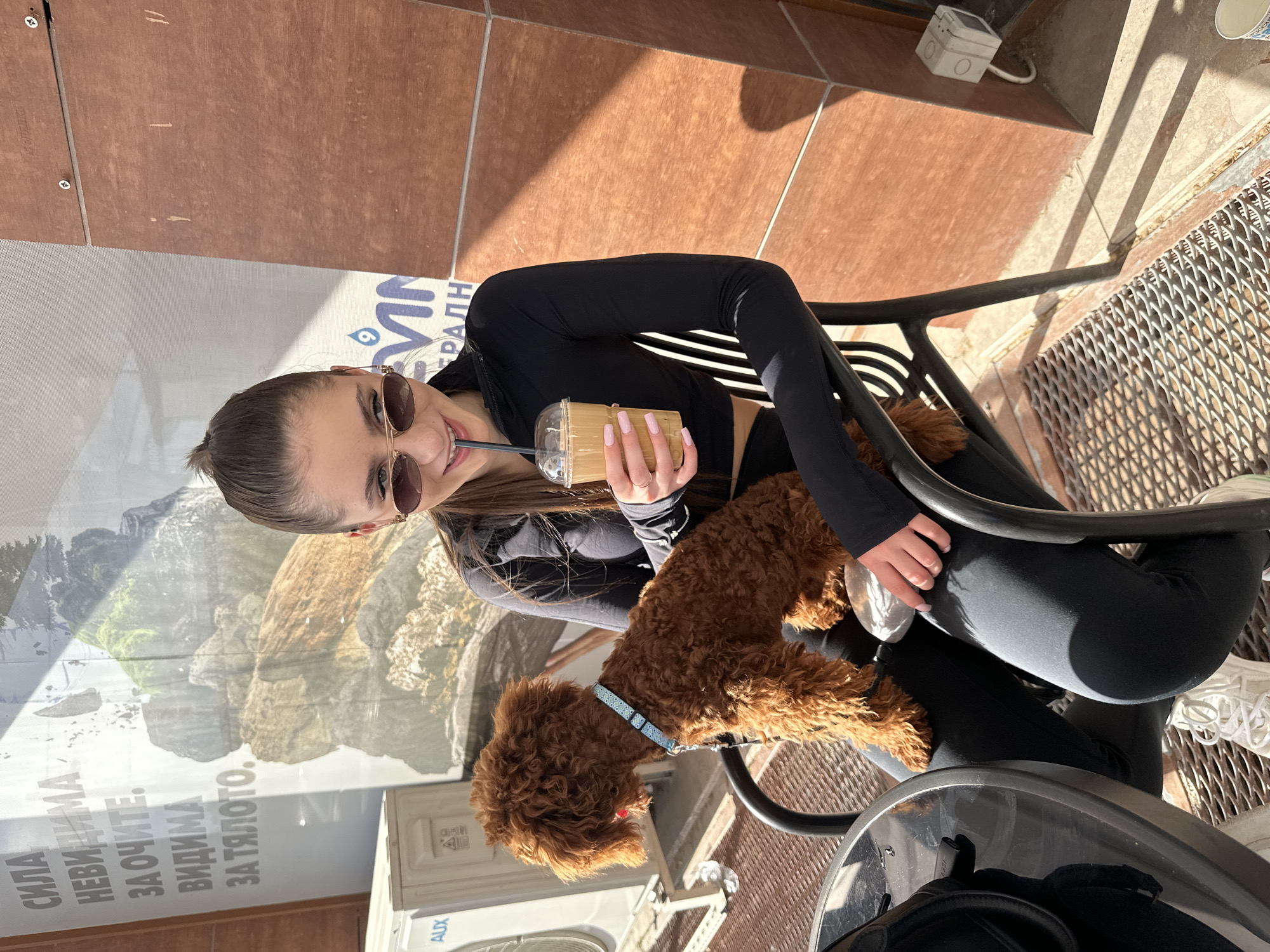 A young woman is captured smiling and relaxing outdoors with her small, fluffy brown dog, likely at an urban cafe or restaurant in Plovdiv, Bulgaria. She has dark hair pulled back, wears sunglasses, a black long-sleeved top, and black leggings, complemented by light pink manicured nails. The woman sips from a clear plastic cup containing an iced beverage with a straw. Her dog, a brown curly-haired breed (possibly a poodle) with a light blue collar, sits on the chair beside her, looking to the side.
The scene is set on a sunny day, with bright daylight casting distinct shadows, suggesting midday or early afternoon. The ground consists of reddish-brown and lighter tiled surfaces, alongside a metal grate. In the background, a large glass panel or window displays a printed image of rocky mountains. Overlaying this image is Cyrillic text, part of an advertisement, with visible words including "СИЛА" (Strength), "НЕВИДИМА" (Invisible), "ЗА ОЧИТЕ" (For the Eyes), "ВИДИМА" (Visible), and "ЗА ТЯЛОТО" (For the Body). A partial logo with a drop icon and the text "VIVA" (or similar) and "ГРАЛН" is also discernable. Below the window, an "AUX" branded air conditioning unit is visible. The woman appears happy and content, enjoying her drink and the company of her pet.