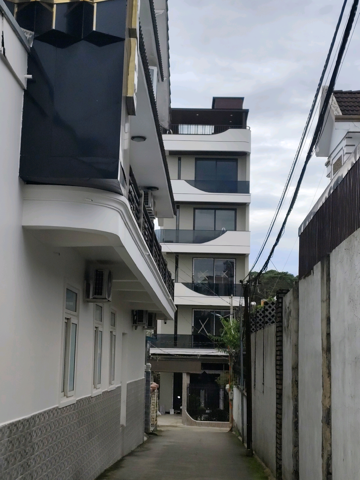 The image depicts a narrow alleyway in Lâm Viên - Đà Lạt, Vietnam, flanked by modern multi-story buildings. The building on the left is white with a textured lower facade and features multiple windows and air conditioning units. The building on the right is taller and has a contemporary design with curved balconies and dark-tinted windows. Overhead, electrical wires are strung across the sky, obscuring a cloudy atmosphere. The alley itself is paved and appears to be a service entrance or a less-trafficked area between properties. The architecture suggests a residential or commercial district with newer developments. There are no people visible in the image.