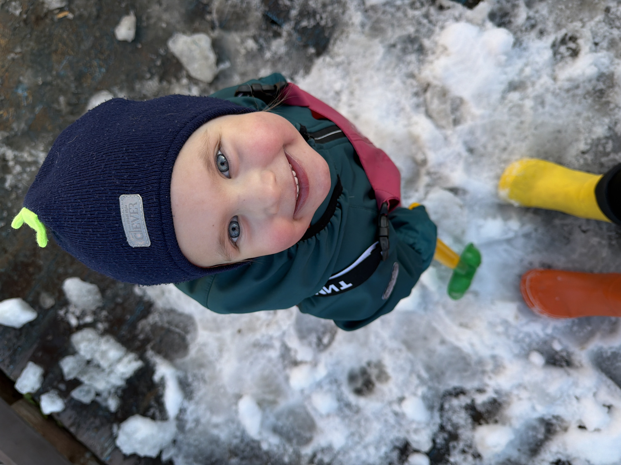 A young child with fair skin and light blue eyes, wearing a dark blue knit hat with a green tassel and a teal winter coat with a maroon lining, looks upwards and smiles. They are standing in a snowy and icy environment. One foot is wearing a bright yellow boot, and another foot is wearing an orange boot. The child appears to be holding a small yellow shovel with a green handle. The surface underfoot is a mixture of snow, ice, and some dark, possibly wet, areas. Based on the presence of snow and the name of the city, this image was likely taken during the winter season in Chaikovsky, Russia.