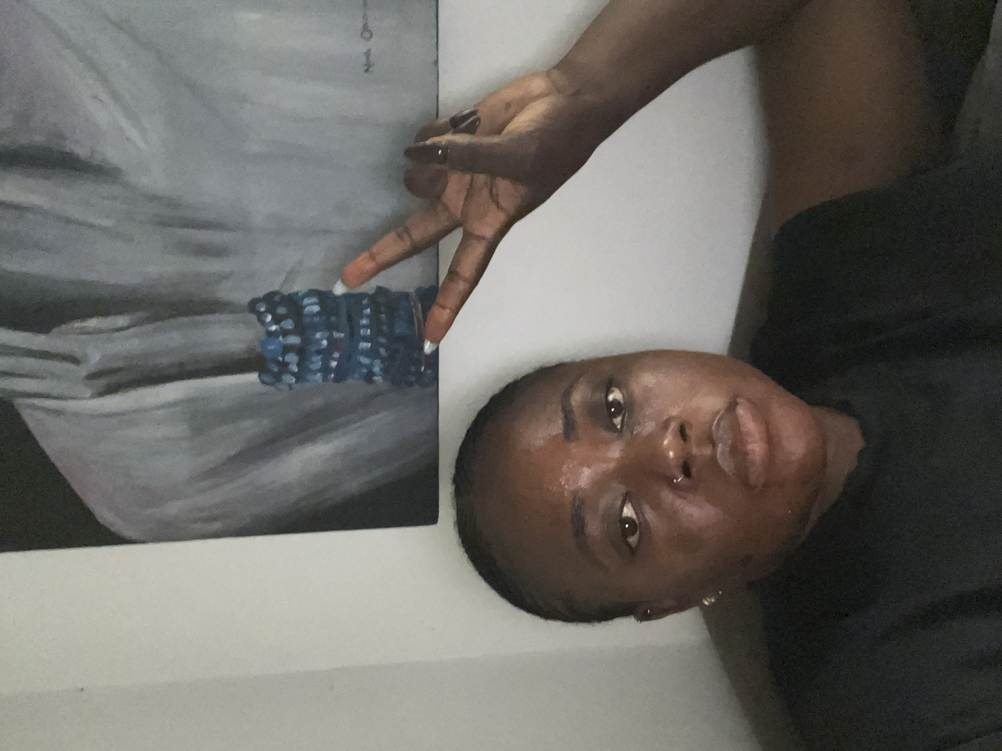 A woman with dark skin and her hair pulled back is looking directly at the camera, her expression neutral. She is wearing a black t-shirt. Her right hand is raised, making a peace sign with two fingers extended. Behind her, on a wall, is a painting. The painting depicts a person wearing a light-colored garment, with a stack of dark blue beaded bracelets placed in front of it. The overall lighting is dim. The location is unknown.
