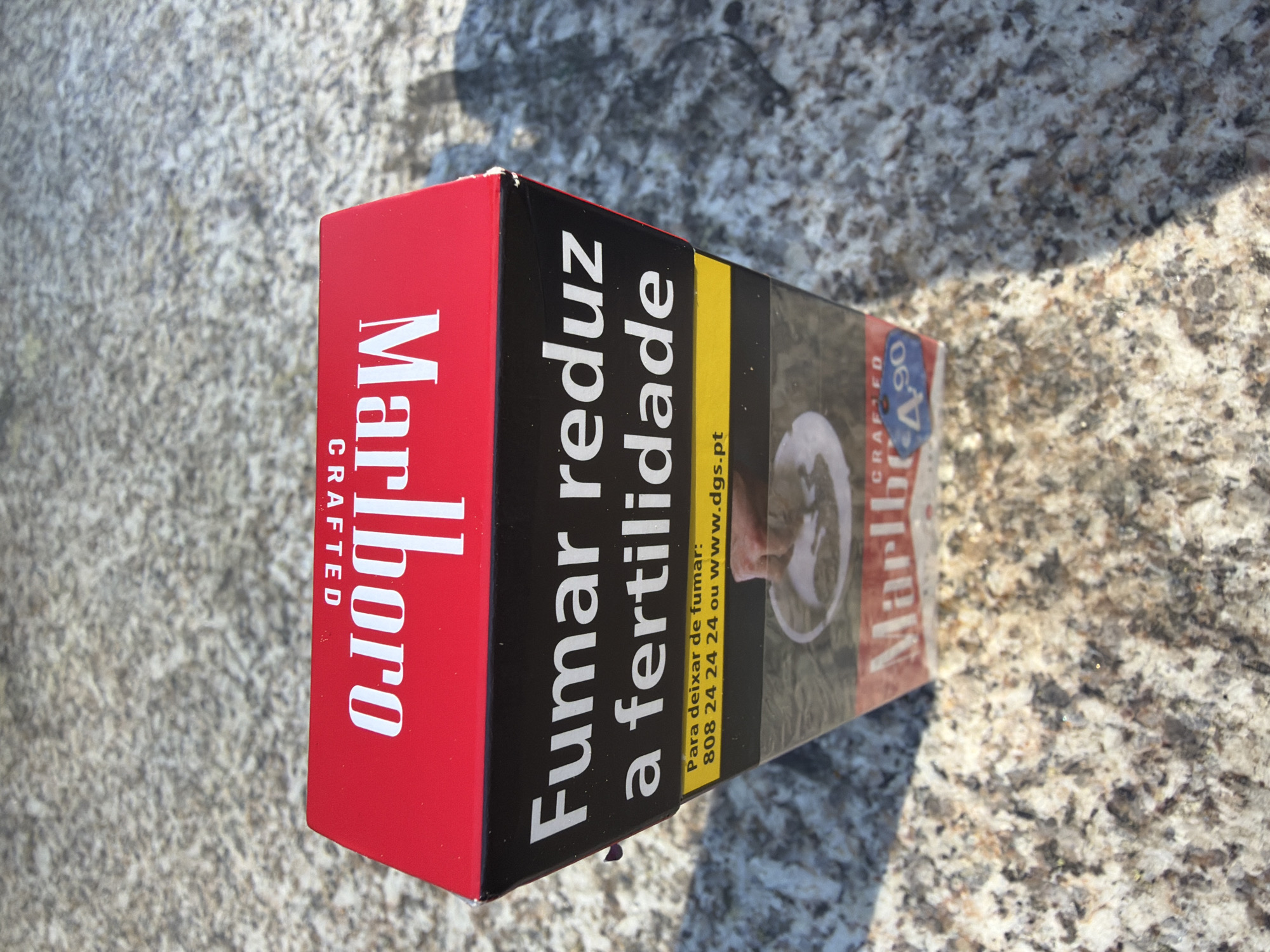 The image shows a pack of Marlboro Crafted cigarettes lying on a mottled gray surface, likely a stone or concrete countertop. The pack is red, white, and black. The text "Marlboro CRAFTED" is prominently displayed on the red side. The adjacent black side features the Portuguese warning "Fumar reduz a fertilidade" (Smoking reduces fertility). Below the warning, on a yellow background, there is text indicating a phone number and website for quitting smoking: "Para deixar de fumar: 808 24 24 24 ou www.dgs.pt". The front of the pack is partially visible, with part of the Marlboro logo and an image of what appears to be a person with a tracheostomy. A blue sticker on the front of the pack is labeled "A 90" and "4,90". The cigarette pack is likely located in Vila Real, Portugal because the warning text is in Portuguese.