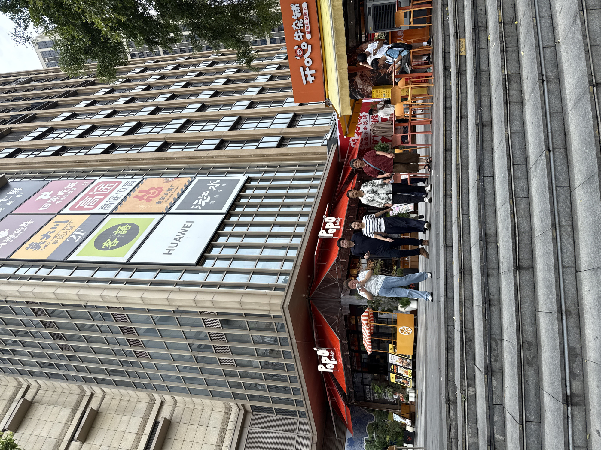 This is a street-level, eye-level shot of a pedestrian area in Haizhu District, China. The foreground consists of gray stone steps leading up to a commercial building. In the midground, several people are walking and standing. On the left, a woman in a white and black striped shirt and dark pants walks forward. Next to her, a woman in a patterned blouse and dark pants walks. To her right, a woman in a maroon shirt and brown pants stands. Behind them, a man in a black shirt and dark pants walks, and to his right, a woman in a white and black striped shirt and dark pants walks.
Further back, under a red awning with "Popo barbar" signage, a food cart is visible. To the right of the awning, an orange building with a yellow awning displays signage in Chinese characters and a cartoon of a bull's head. A person in a white shirt and shorts is seated at a table on the right side, looking at a phone. A person in a blue t-shirt and dark shorts is also seated at a table to the right, looking at a phone.
The background is dominated by a tall, modern building with a facade of glass windows and concrete or stone elements. A large sign with multiple panels featuring Chinese characters and logos, including "HUAWEI," is attached to the building. Trees and another building are visible in the distance. The sky is overcast. The overall impression is a bustling urban commercial area.