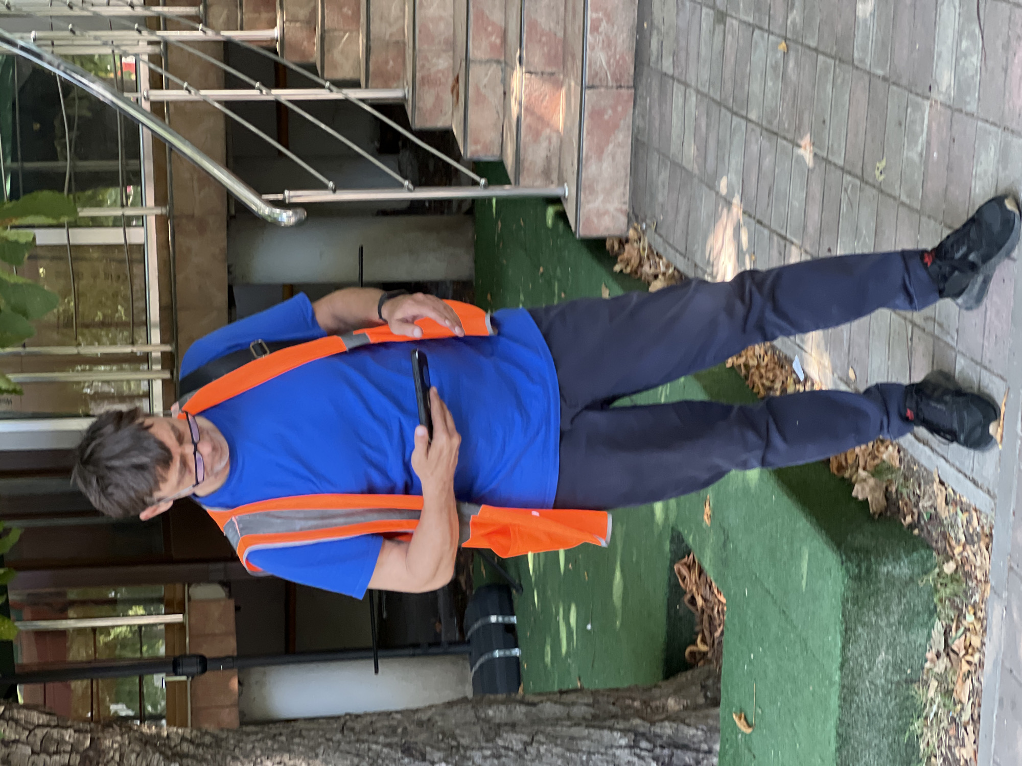 An adult male wearing a bright blue t-shirt, an orange safety vest, and dark pants stands on a paved sidewalk next to a green-covered raised area. He is looking down at a black smartphone in his hands. The man has short brown hair and is wearing glasses. Behind him, a metal railing leads up a set of outdoor stairs made of brick and stone. A tree trunk is visible to the left of the man. The scene appears to be outdoors in a public area, possibly a park or commercial district, in Krasnodar, Russia.