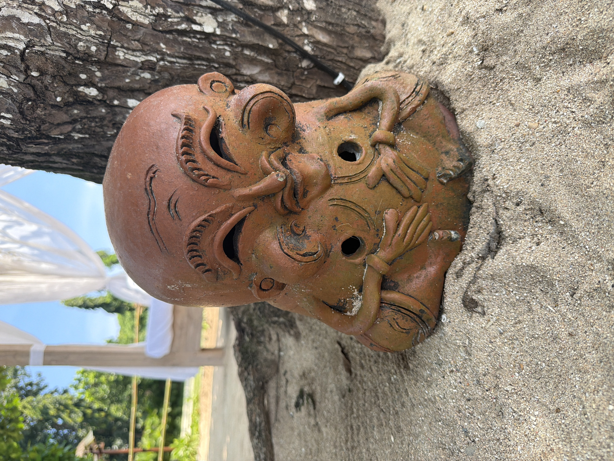 A weathered, terracotta-colored earthenware statue of approximately 30-40 centimeters in height is depicted partially embedded in light brown, coarse sand. The statue features a stylized, grotesque-like human face with a bald head, smiling eyes with raised eyebrows, an exaggerated nose, and prominent cheeks. Below the main facial features, two circular holes are present, flanked by sculpted arms and hands holding the lower portion of the figure. The statue's surface exhibits erosion, sand particles, and patches of light green and white discoloration.
In the mid-ground to the right, a section of a mature tree trunk with deeply furrowed, dark bark and white lichen patches is visible directly behind the statue. A black electrical cable runs vertically down the tree trunk. To the left and slightly further back, a light-colored wooden four-poster daybed with a white mattress and draped white sheer fabric is positioned. The background consists of blurred, dense green tropical foliage and trees under a clear, bright blue sky. The scene is illuminated by daylight. This location is identified as a beach or resort area within Nusa Dua, Indonesia.