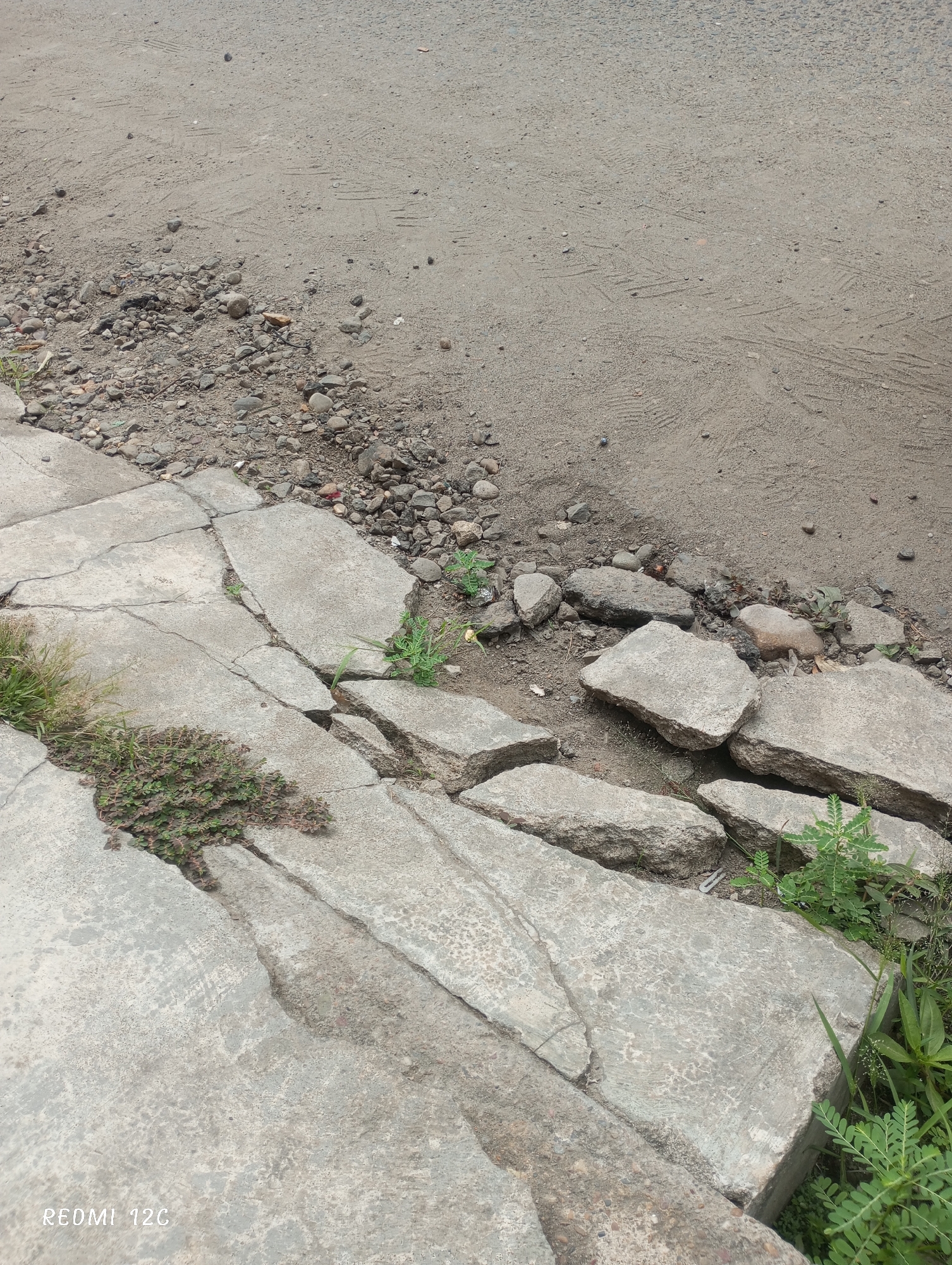 The image shows a broken and cracked concrete sidewalk in Lhokseumawe, Indonesia. The sidewalk edges are crumbled, with some fragments displaced and surrounded by dirt and small stones. Weeds and grass are growing through the cracks in the concrete. The sidewalk abuts a dirt or sandy surface, which extends into the upper portion of the image. The lower left corner of the image contains the text "REDMI 12C."