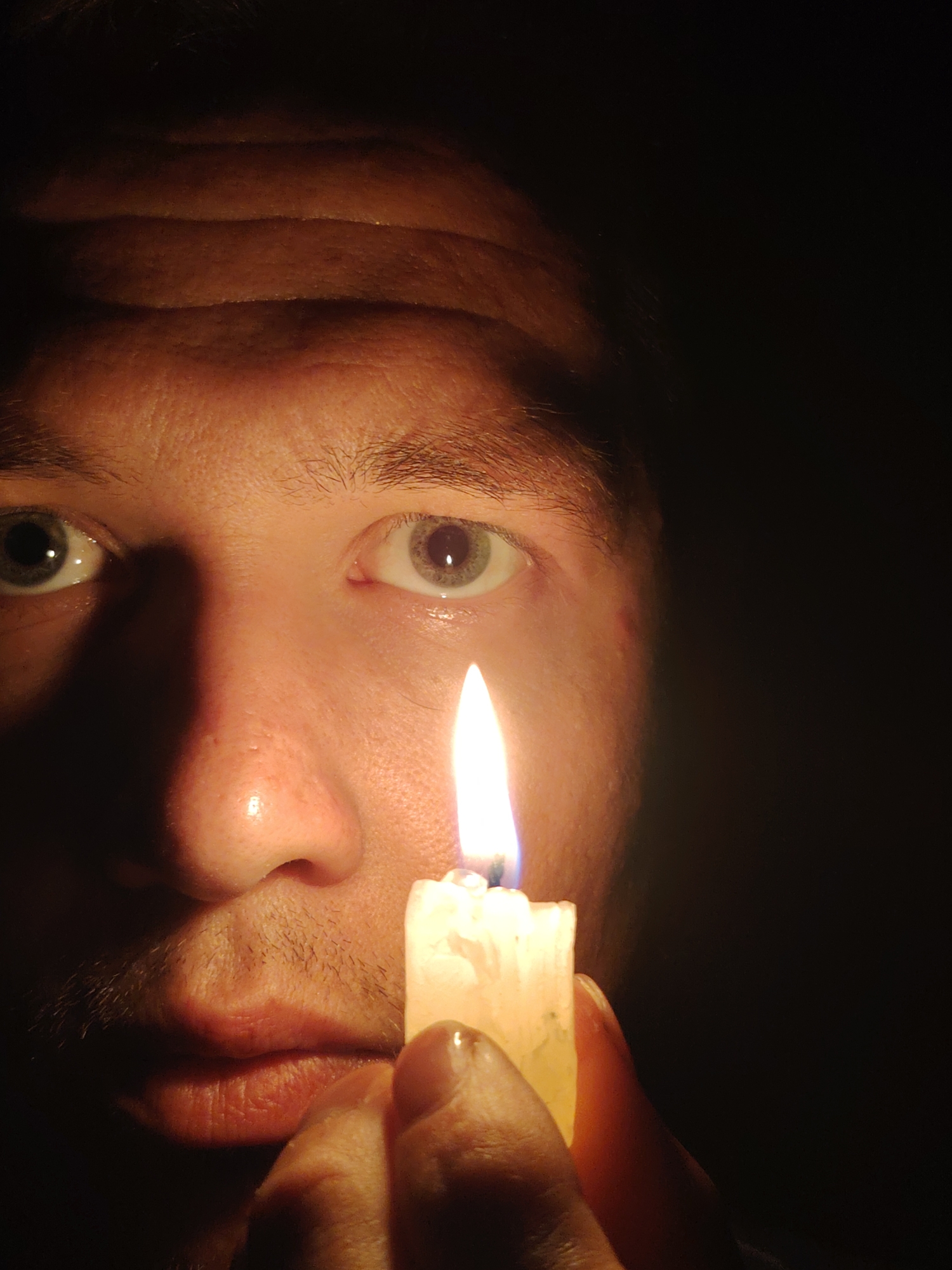 A close-up, low-angle shot shows the face of a person illuminated by a candle. The person's forehead is furrowed and their eyes are wide open, reflecting the flame. A lit candle is held close to the person's face, casting dramatic shadows. The background is dark, obscuring any details of the surroundings. The image is cropped to focus on the face and the candle, suggesting an intimate or perhaps unsettling atmosphere. Without further context, it is impossible to determine the exact location, but the setting appears to be indoors in a dimly lit environment, possibly a home. There are no specific landmarks or details to place this image in Chaikovsky, Russia.