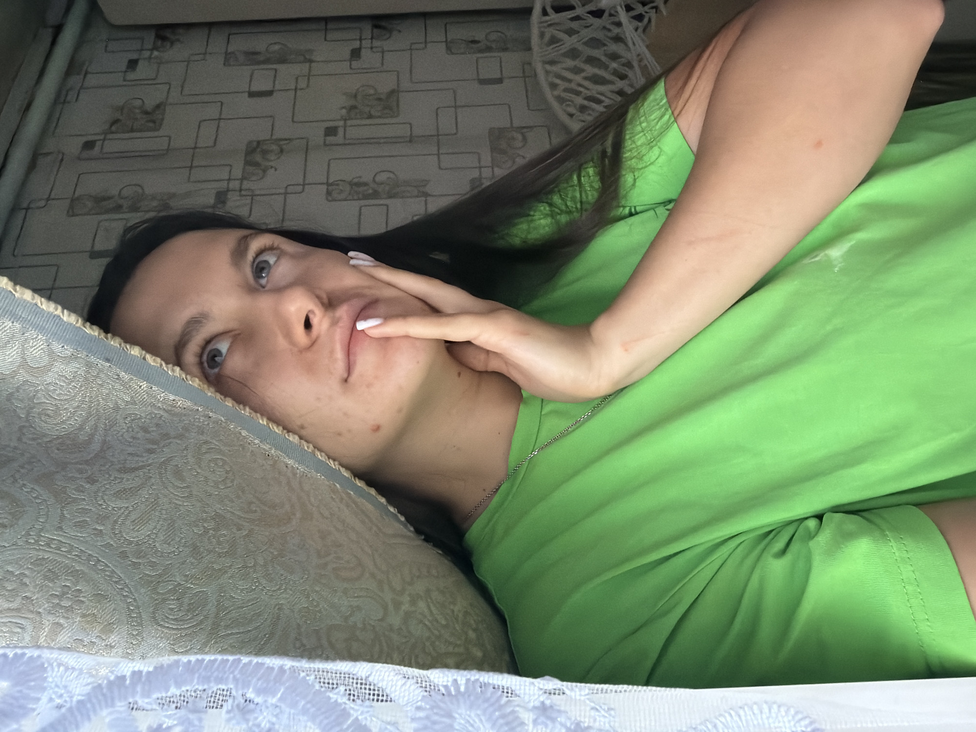 A young woman with dark hair and blue eyes is pictured from the chest up, looking upwards and to her right. She is wearing a bright green t-shirt and has her right hand touching her face. A portion of a curtain is visible in the upper left corner of the image, with a light blue lace trim. The background appears to be an interior wall with a geometric pattern. The lighting suggests an indoor environment, possibly a room in a building located in Красный Маяк, Russia.