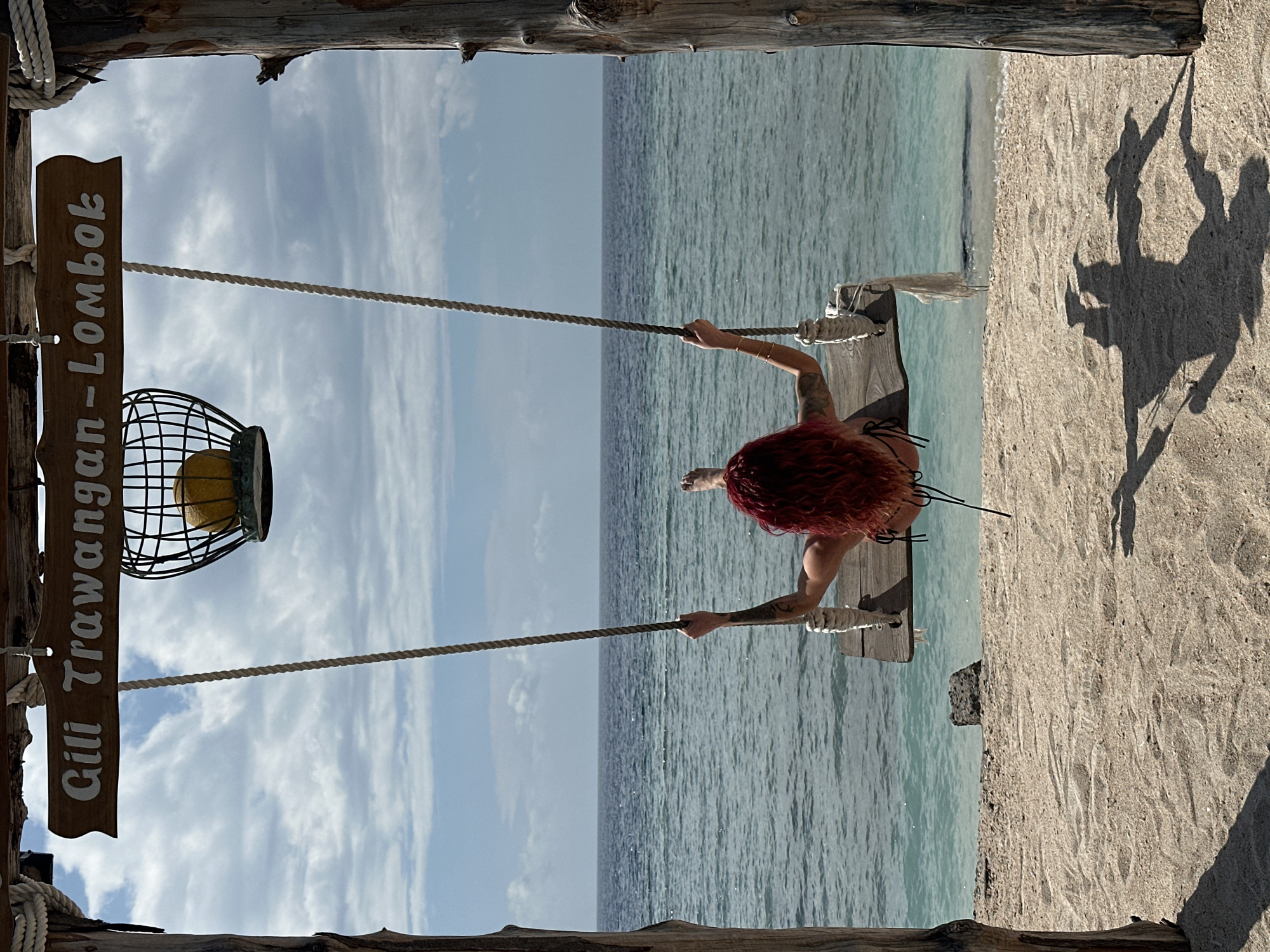 A woman with red hair is swinging on a swing over a body of water at Gili Trawangan, Lombok, Indonesia. The swing is suspended by ropes and attached to a wooden frame. The woman is facing the water and her shadow is visible on the sandy beach. A sign with the words "Gili Trawangan - Lombok" is attached to the wooden frame. The sky is partly cloudy.