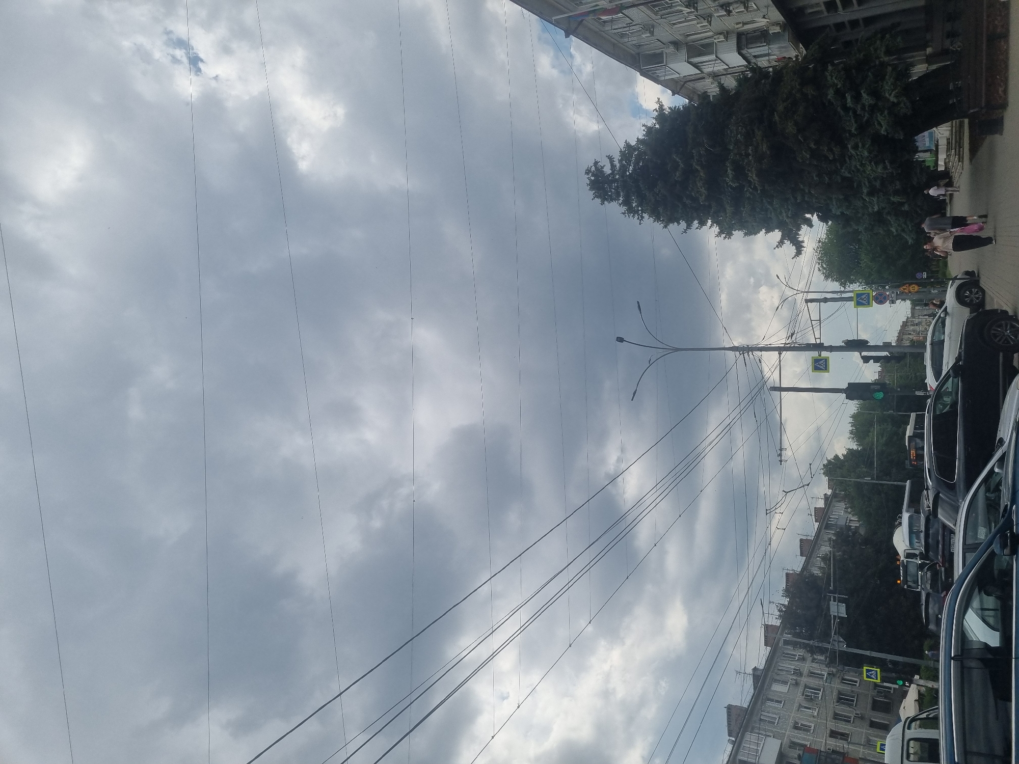 This image captures a street scene in Krasnodar, Russia, under an overcast sky. The perspective is from a moving vehicle, looking upwards and towards the right. The primary subjects are the buildings lining the street, a large evergreen tree, overhead power lines, traffic lights, and several vehicles.
The buildings appear to be multi-story residential blocks with numerous windows, characteristic of urban Russian architecture. The evergreen tree is dense and dark green, providing a stark contrast to the muted colors of the sky and buildings. Several cars, including a white car in the foreground and a black SUV, are visible, indicating a city street with traffic. A white van is also partially visible in the lower right corner.
Overhead, a complex network of power lines crisscrosses the frame, suggesting the area is serviced by electricity and possibly public transport like trams or trolleybuses, though these are not directly visible. Traffic lights are present, with one showing a green light, implying the flow of traffic. Pedestrians, two women in the middle ground, are walking along the sidewalk near the tree and buildings.
The weather appears to be cloudy, with the sky filled with a mix of light and dark gray clouds. There is no strong sunlight, suggesting it could be morning or late afternoon, or simply a cloudy day. The overall mood is calm and ordinary, typical of a day in a city. There is no discernible text in the image, except for what might be on street signs or license plates which are too small to read. The scene depicts a normal urban environment in Krasnodar.
