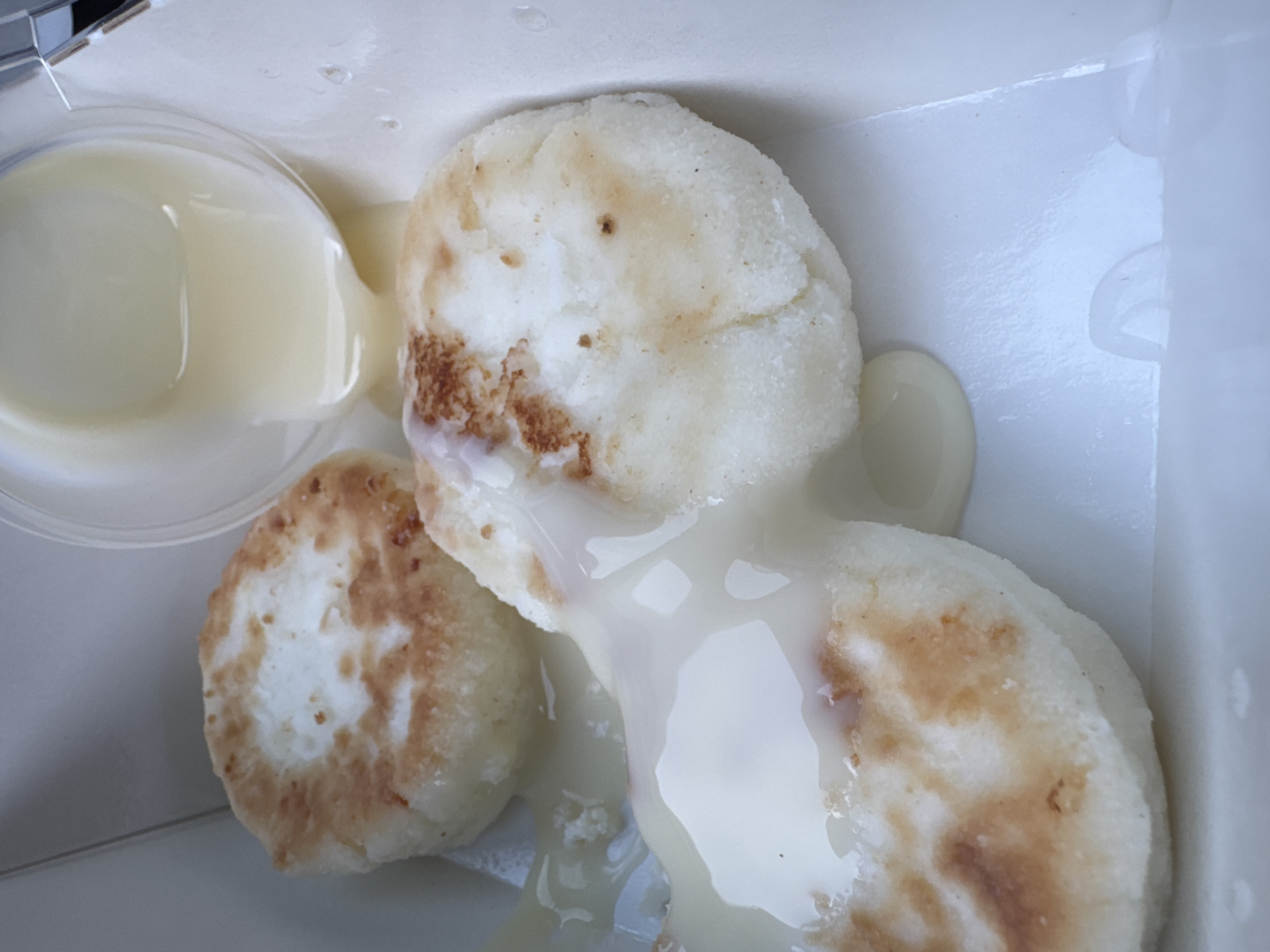 A close-up, overhead shot displays three syrniki (small cheese pancakes) drenched in a white sauce inside a white container. A small, clear plastic cup filled with the same white sauce sits to the left of the pancakes. The pancakes are round, lightly browned, and slightly uneven in shape. The white sauce appears creamy and glossy. The container appears to be a takeout box. The image was taken in Chaikovsky, Russia.