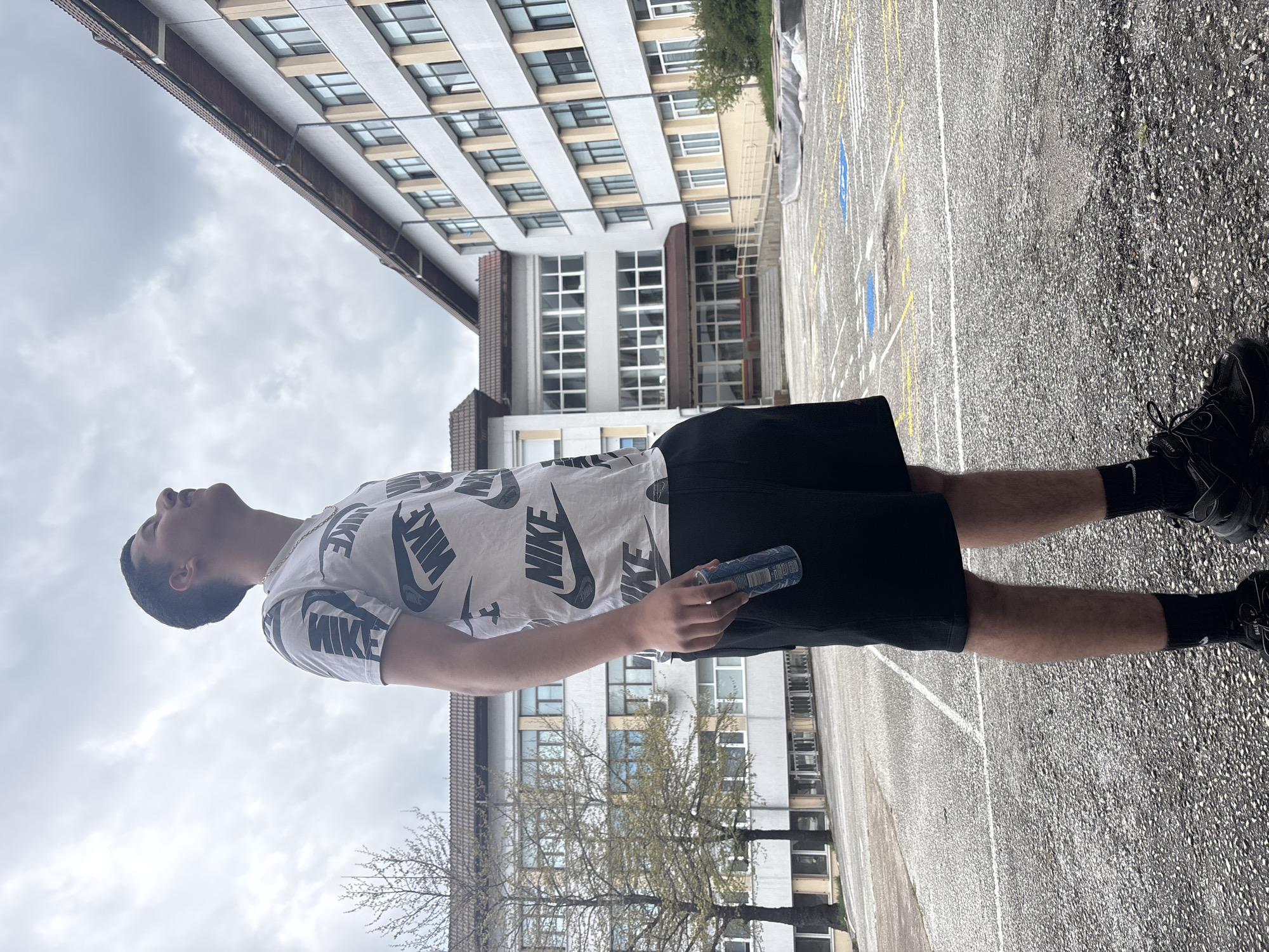 A young man stands in what appears to be a parking lot or paved area, possibly in Zlatograd, Bulgaria given the prompt. He is wearing a white t-shirt with "NIKE" printed repeatedly in black, black shorts, and black sneakers with black socks. He is holding a cylindrical blue can in his right hand. He is looking upwards towards the sky. A large building with multiple windows is behind him. The sky is overcast. White and yellow lines are painted on the pavement of the parking lot. A tree with sparse foliage is also visible.