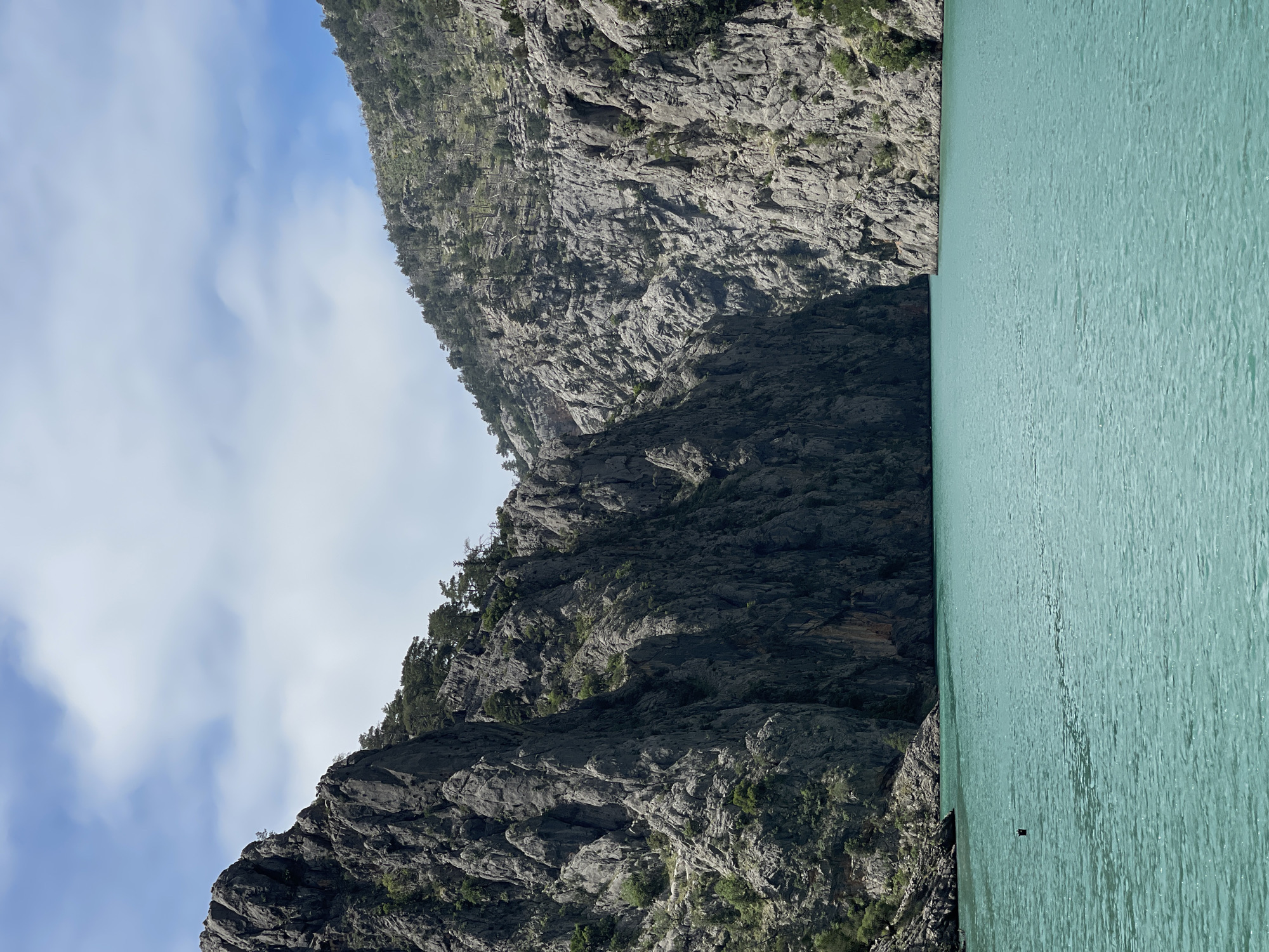 The image presents a scene of mountainous terrain adjacent to a body of water, possibly a lake or fjord. The mountains are rugged, with visible rock formations and sparse vegetation. The water is light green, suggesting the presence of minerals or algae. The sky is partially visible, showing a light blue color with some cloud cover. The location is Unknown, Unknown.