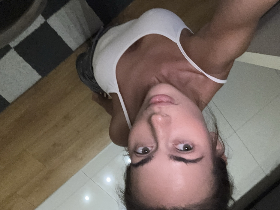 A woman takes a selfie from a low angle, tilting the camera upwards. She is wearing a white tank top and dark-colored shorts. The background includes a wooden floor, a black and white checkered rug, and a glimpse of a light-colored wall. Her hair is pulled back, and her eyes are looking directly into the camera.