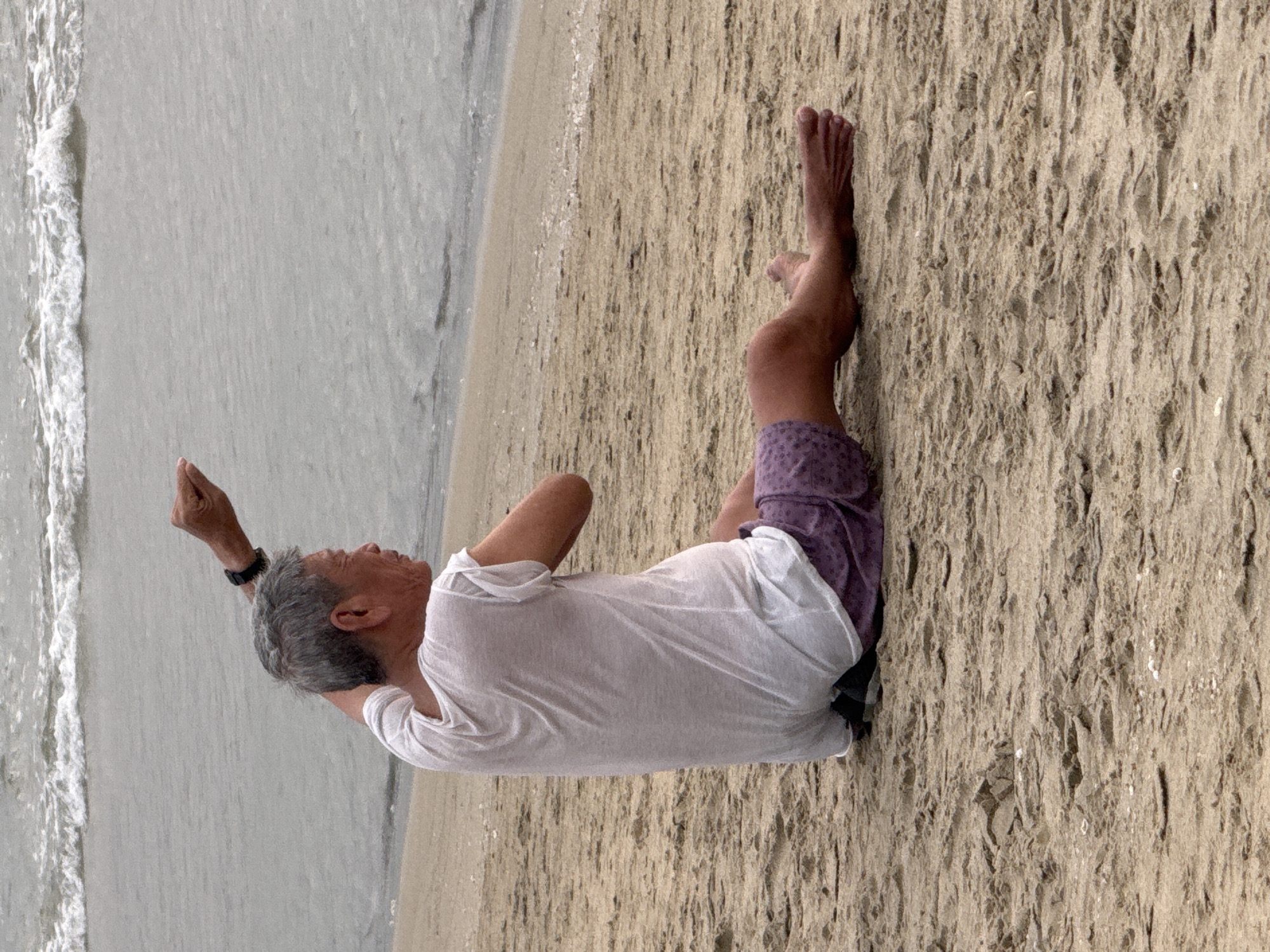 A man in a white T-shirt and purple shorts is lying on the sand at a beach. He is on his back, facing the ocean, with his right arm raised. The water is gray, and there are waves washing ashore. Based on the location, this could be one of the beaches in Đà Nẵng, Vietnam.