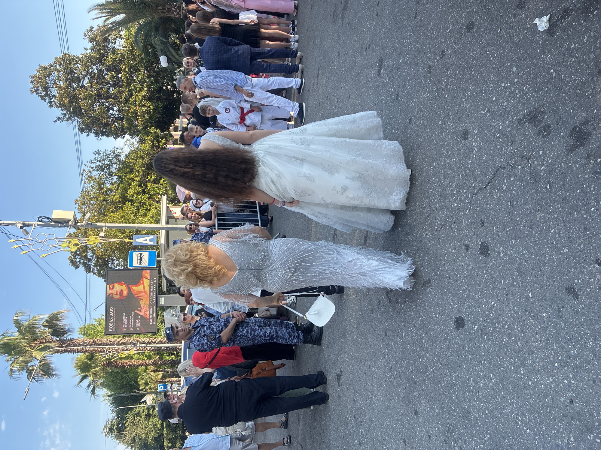 In Sochi, Russia, at what appears to be an event on a street with palm trees and lush vegetation, a group of people are gathered. Two women are in the foreground, one in a long, sparkling silver gown with feathered details and the other in a white, lace-adorned ball gown. The woman in the silver dress is holding a white purse. Behind them, a diverse crowd is visible, including individuals in casual attire, a young boy in a white karate gi with a red belt, and people in what appear to be security or law enforcement uniforms. A large digital billboard displays an advertisement with text in Russian. The ground is asphalt, with some dark stains and debris. The scene suggests a public gathering or celebration, possibly an outdoor event or promenade.