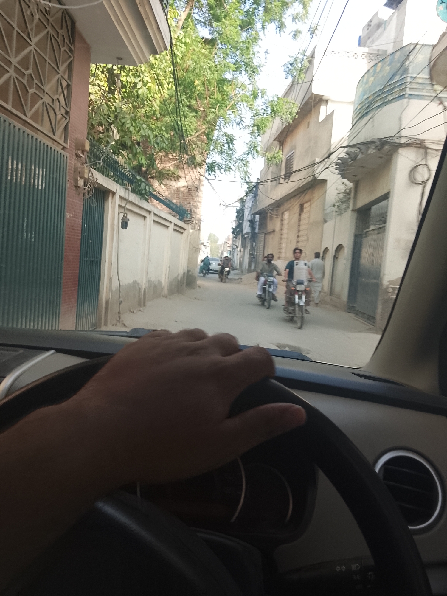 The image depicts a view from inside a vehicle, showing a street scene in an urban area of Unknown, Unknown. A driver's hand is visible on the steering wheel. In the street ahead, several people are riding motorcycles and walking. Buildings line both sides of the street, with various gates and walls visible. Overhead power lines crisscross above the street. Trees are interspersed among the buildings, adding some greenery to the scene.