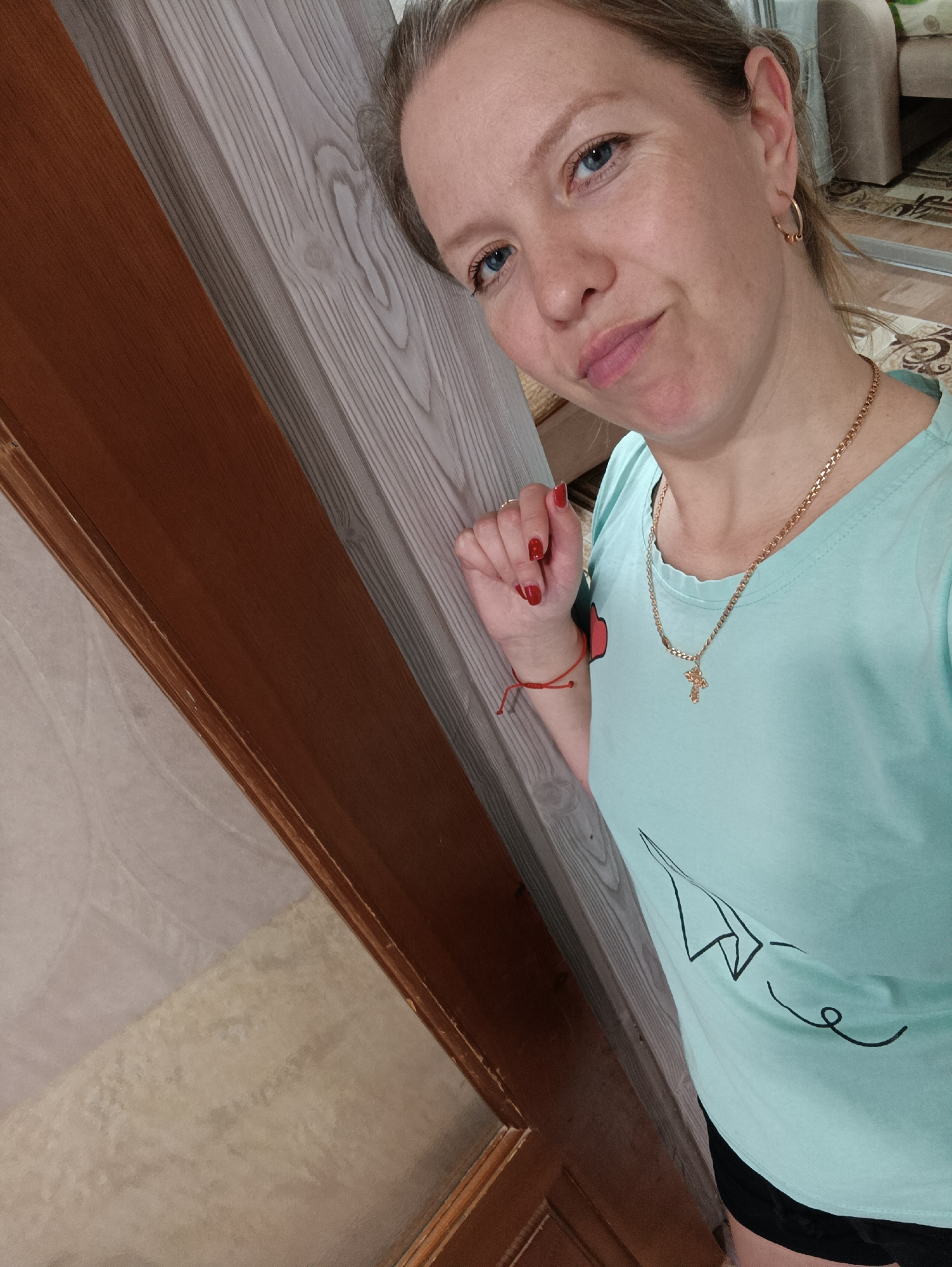 A woman with fair skin and blue eyes is pictured in an indoor setting, possibly a home. She is wearing a light blue T-shirt with a paper airplane graphic and a gold cross necklace. Her nails are painted red, and she has small gold hoop earrings. She is leaning against a wooden door frame with a lighter-colored panel. The door is partially visible, with a frosted glass panel. In the background, there is a glimpse of furniture and a patterned carpet. The location might be in the city of Chaikovsky, Russia, given the user's prompt.