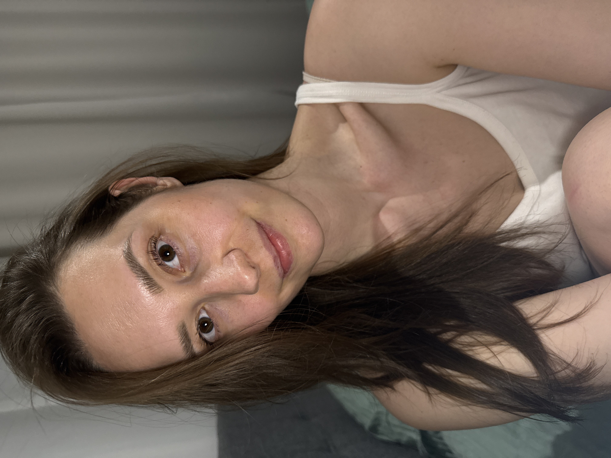 A young adult female with long, straight, dark brown hair and fair skin is depicted in a close-up, frontal portrait. She has brown eyes and a subtle smile. She is wearing a white ribbed tank top with thin straps. Her right shoulder, upper chest, and part of her right knee are visible, suggesting a seated or reclining posture. The background is out of focus, revealing a dark grey/teal-colored pillow or fabric on the left and a light grey/off-white curtain or vertically textured wall on the right. Lighting appears soft and even, originating from the front. The specific location within the city of Chaikovsky, Russia, cannot be determined from the visual information provided; the scene is an indoor domestic setting.