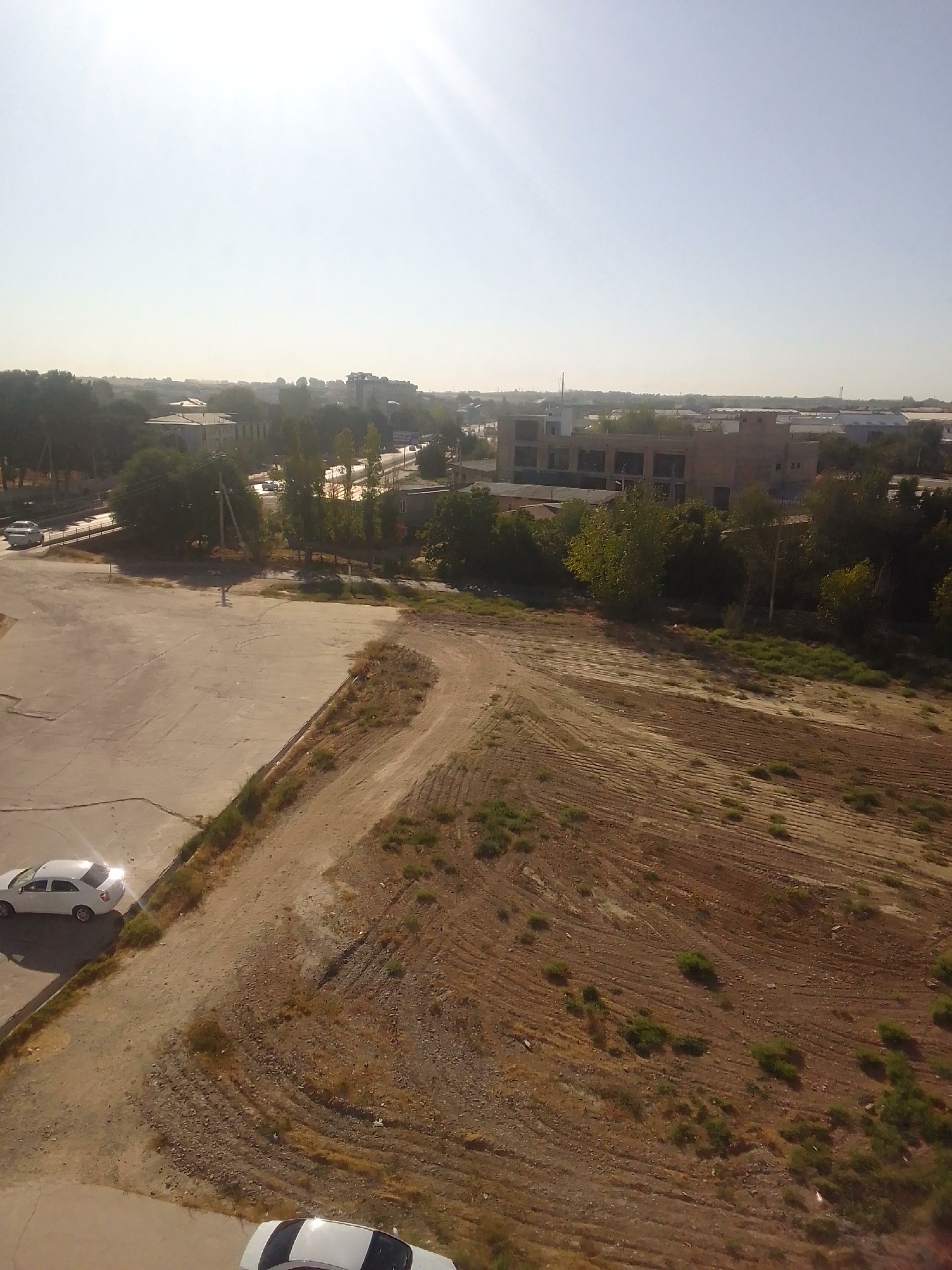 The image depicts an outdoor scene in Kattakurgan City, Uzbekistan, likely during daytime given the bright sunlight. In the foreground, a section of a paved area, possibly a parking lot or road, is visible. A white car is parked on this paved surface. Adjacent to the paved area is a section of dry, brown earth with visible tracks or furrows, suggesting it might be a field or undeveloped land. Scattered patches of green vegetation are present on the earthy ground.
In the middle ground and background, a road with some traffic can be observed, bordered by trees. Further in the distance, buildings and structures, including what appears to be an unfinished or industrial building with multiple windows, are visible. The sky is clear and bright, with the sun prominently featured, causing lens flare in the upper portion of the image. The overall impression is of an urban or semi-urban environment with elements of construction and nature.
