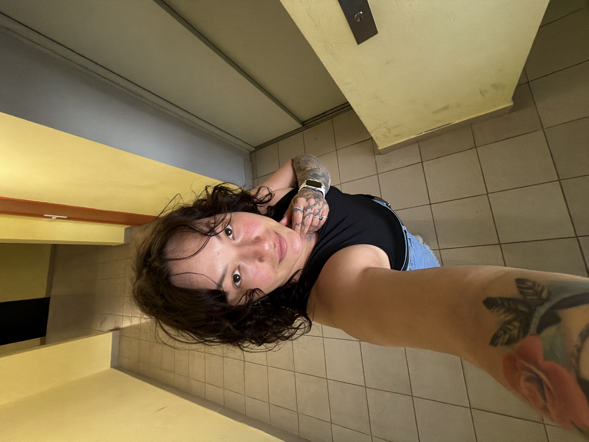 A woman with dark, curly hair and tattoos is standing in a tiled hallway with yellow walls, taking a selfie. She is wearing a black tank top and denim shorts. To her right is a large, grey elevator door. The hallway appears to be in an apartment building in the city of Chaikovsky, Russia.
