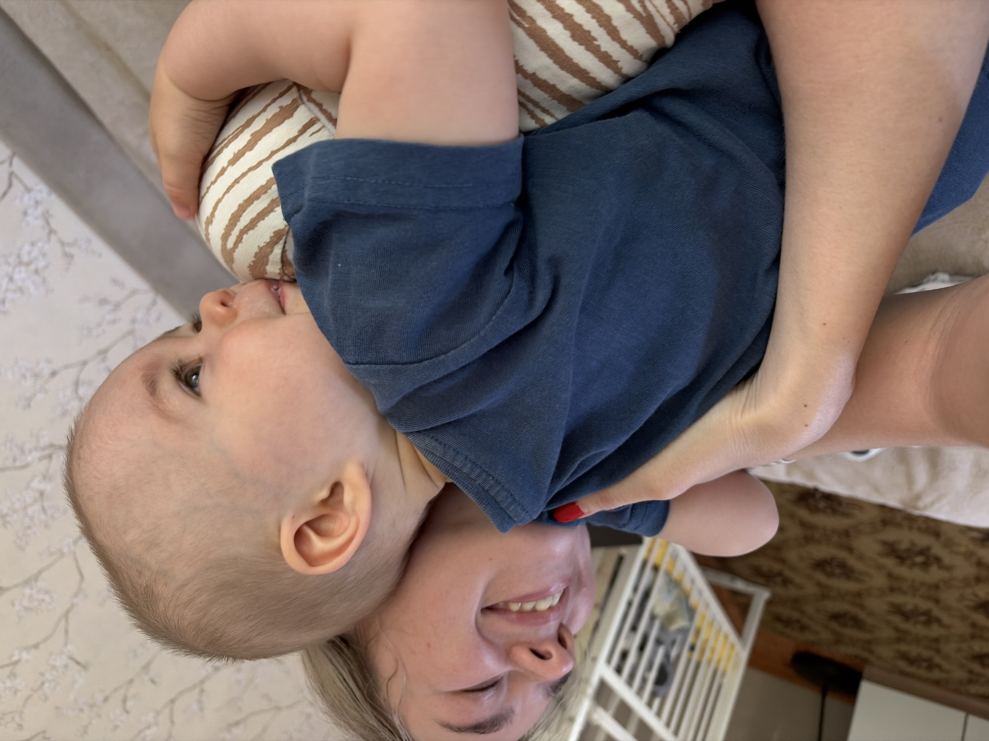 A woman is lying on her back, holding a baby. The baby is wearing a blue shirt and is facing to the left. The woman has blonde hair and is smiling. The baby is looking up at something to the left of the frame. A patterned pillow is under the baby's head. A crib with white and yellow railings is visible behind the woman. The image appears to have been taken indoors. The location is inferred to be in Chaikovsky, Russia.