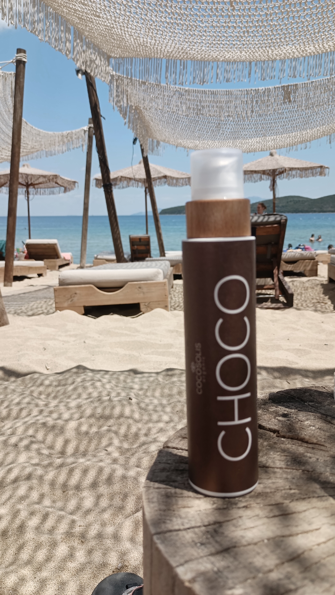 The image depicts a beach scene in Nea Peramos, Greece, on a sunny day. In the foreground, a brown bottle with a white label is placed on a wooden surface. The bottle has the word "CHOCO" printed vertically in large white letters and "cocosous" printed vertically in smaller white letters. Behind the bottle, the sandy beach is visible, with shadows cast by overhead fabric canopies. Further in the background, there are beach chairs, umbrellas, and the sea. A few people are visible near the water. The overall atmosphere is relaxed and summery.