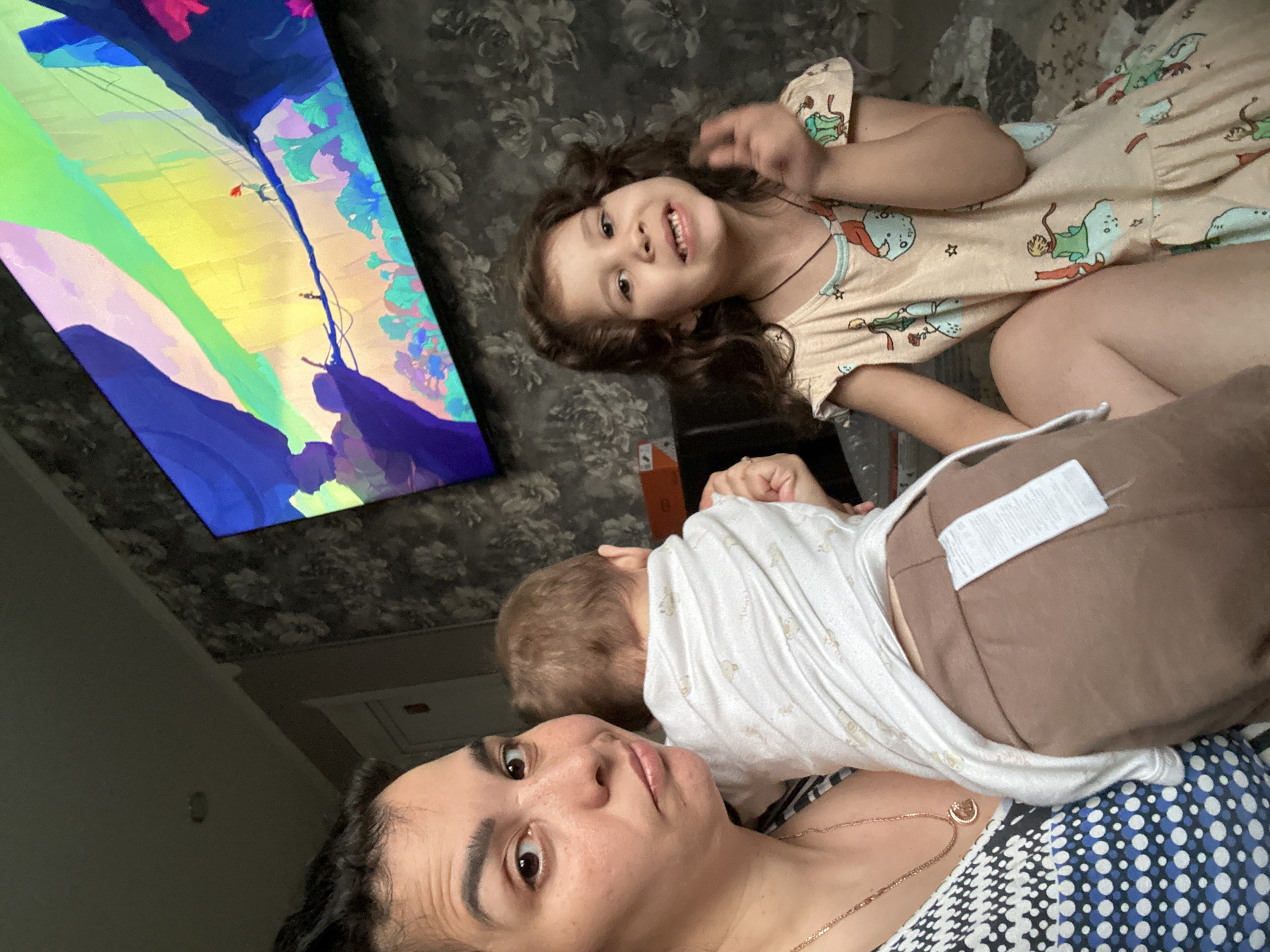 This image captures a relaxed indoor family scene featuring a woman and two children. A woman lies on her back, looking at the camera with a neutral expression. She has dark hair and wears a patterned top with a thin gold necklace. A baby or young toddler with light brown hair is resting on her, seen from behind, wearing a white top and brown bottoms with a white velcro-like patch.
Beside them, a young girl with curly dark hair smiles cheerfully at the camera, her hand slightly raised. She wears a light-colored dress adorned with various cartoon characters.
In the background, a large television screen displays a vivid, abstract image or animation characterized by bold blocks of blue, green, yellow, and purple. The walls are covered with dark, patterned wallpaper, possibly featuring floral designs, suggesting a cozy home environment. The scene depicts a casual family moment, likely during the daytime, as they relax together, possibly watching television. No visible text is present in the image.