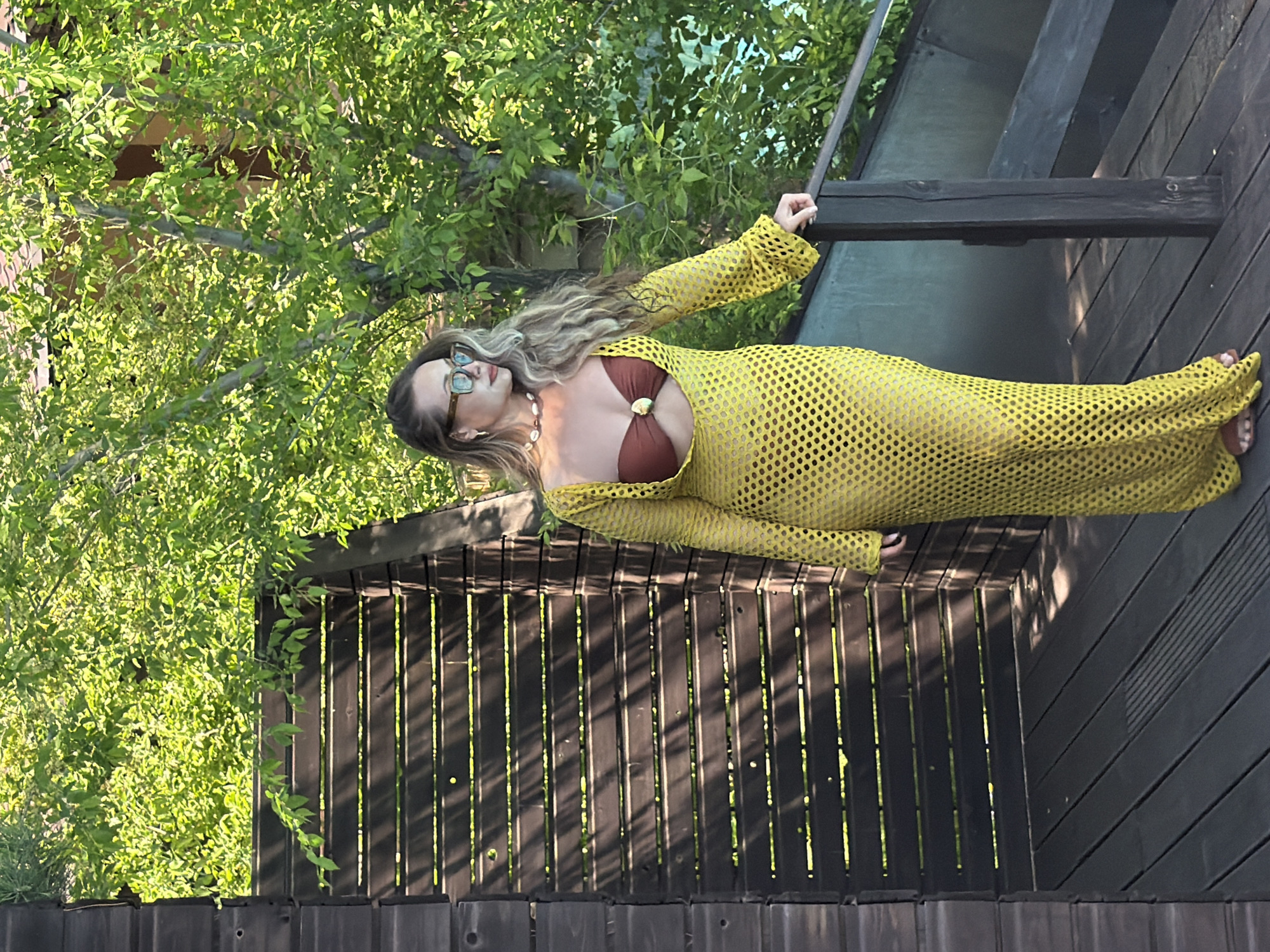A woman with long, wavy blonde hair and glasses poses outdoors, looking to her right. She is wearing a bright yellow, long-sleeved, netted cover-up over a brown bikini top, which features a golden, shell-like embellishment. A multi-strand shell necklace is visible around her neck. Her right arm rests on a dark wooden railing.
The setting appears to be a wooden deck or balcony, characterized by dark wooden planks and vertical railings that cast distinct shadows. Behind the woman, lush, vibrant green foliage, likely from trees or bushes, fills the background, suggesting a natural and verdant environment. The scene is illuminated by bright daylight, indicating a clear, sunny day, with strong sunlight highlighting the leaves and creating distinct shadows on the wooden structures. No text is visible in the image. The location context provided is Krasnoyarsk, Russia.