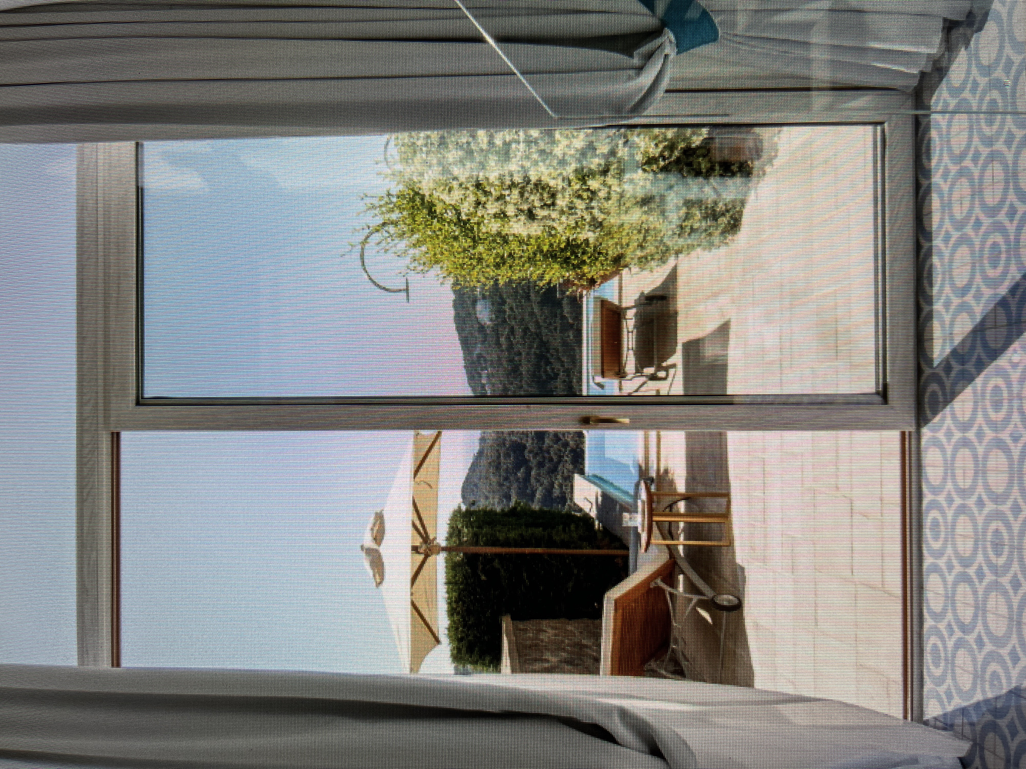 This is a photograph taken from inside a room looking out through a sliding glass door. The view is of a patio area with a swimming pool, lounge chairs, and an umbrella. Beyond the patio, there are rolling hills covered in trees, suggesting a natural or semi-rural setting. The foreground shows a patterned tiled floor and the edge of a curtain. There are no people visible in the image. The scene depicts a tranquil outdoor space with a view of nature. This location is not in Łódź, Poland.
