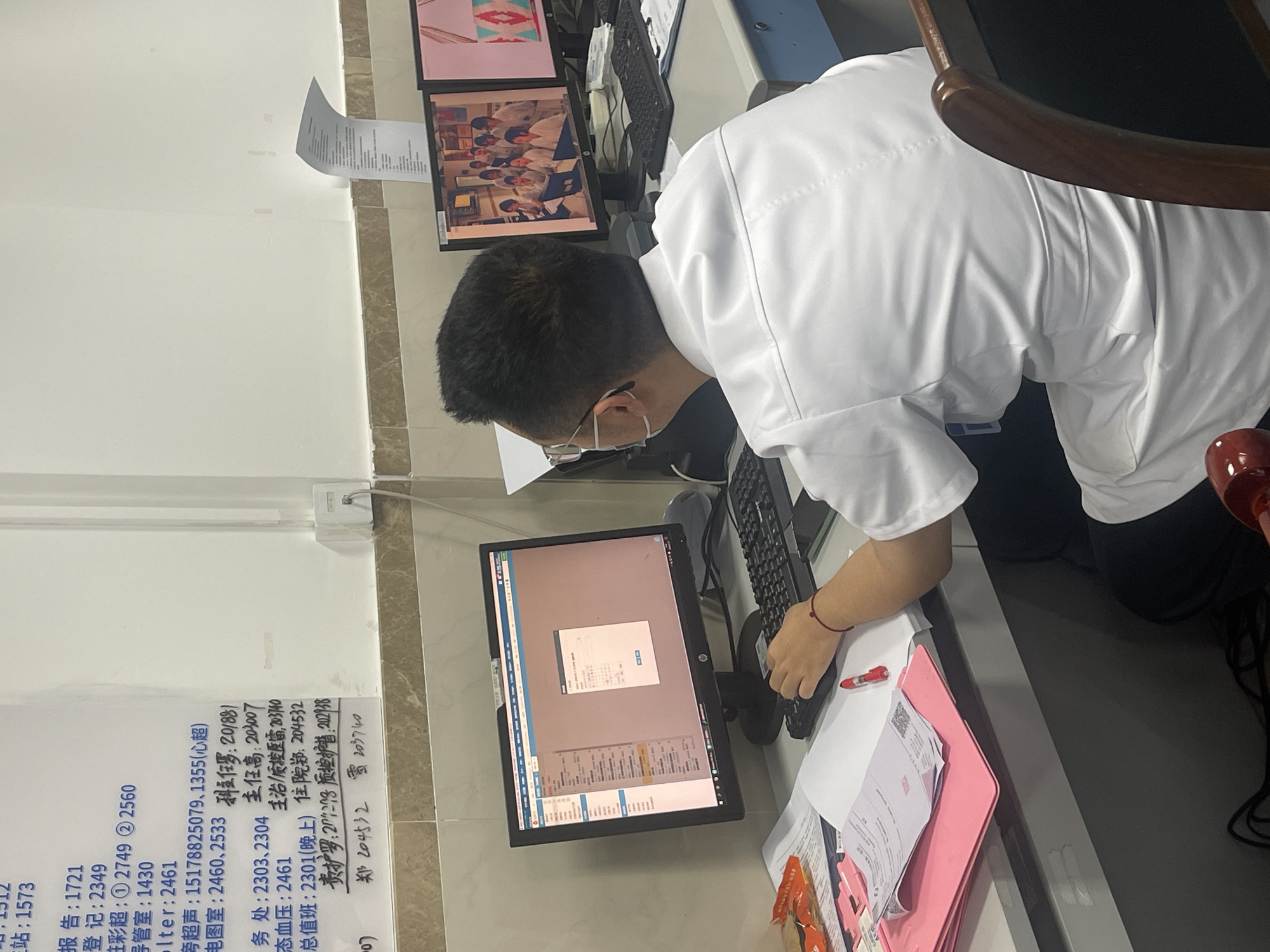 A person wearing a white shirt and glasses is sitting at a desk in what appears to be an office or medical setting. The person is using a computer and a keyboard, surrounded by papers and files. Additional computer monitors are visible in the background. A wall with a list of numbers written on it is in the upper-left. Based on the Chinese characters on the wall, the location may be Yuzhong District, China.