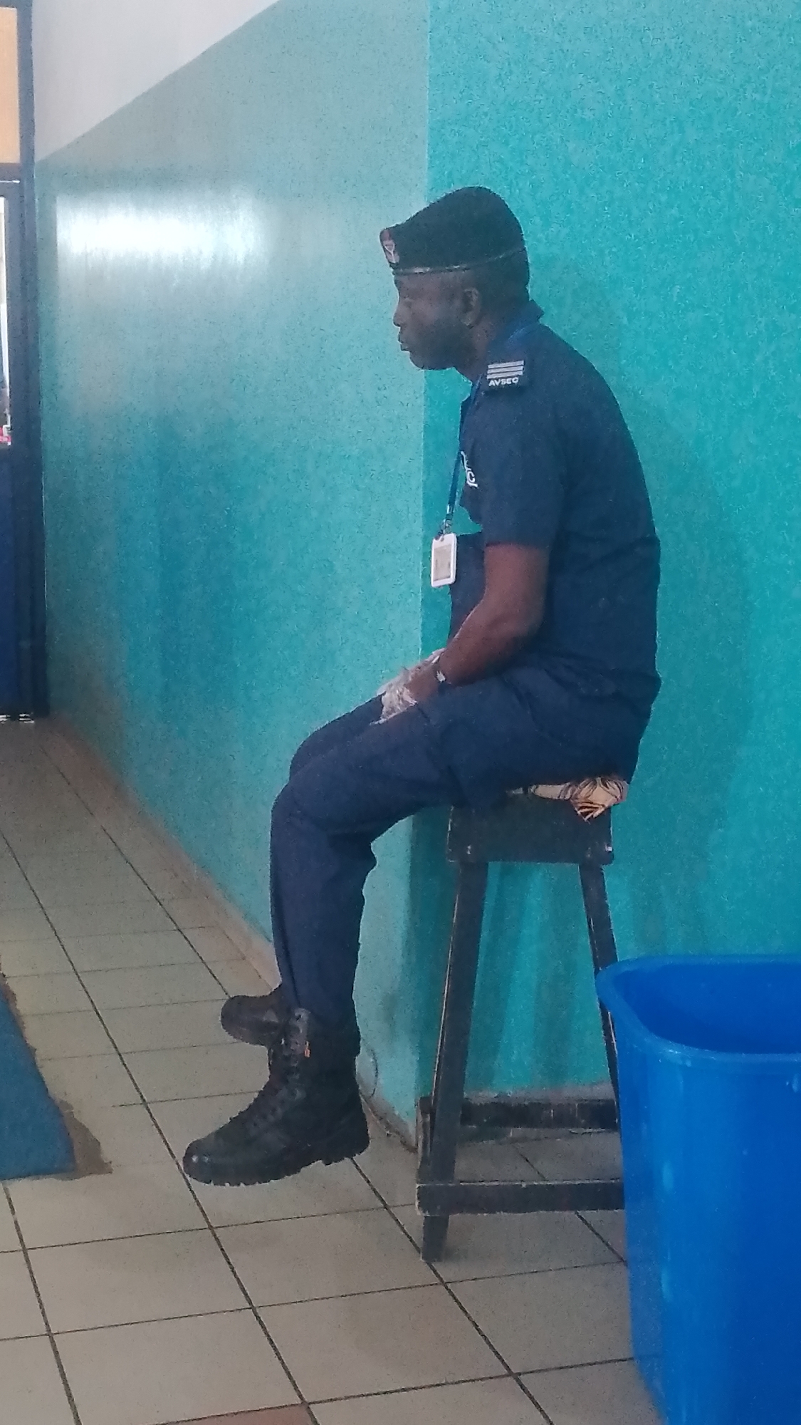 A man wearing a dark blue uniform and a black beret is seated on a stool. He has a white ID card hanging around his neck. He is wearing black boots. He is in a room with a turquoise wall. A blue plastic trash bin is beside the stool. The floor is tiled. The location is identified as Orerokpe, Nigeria.
