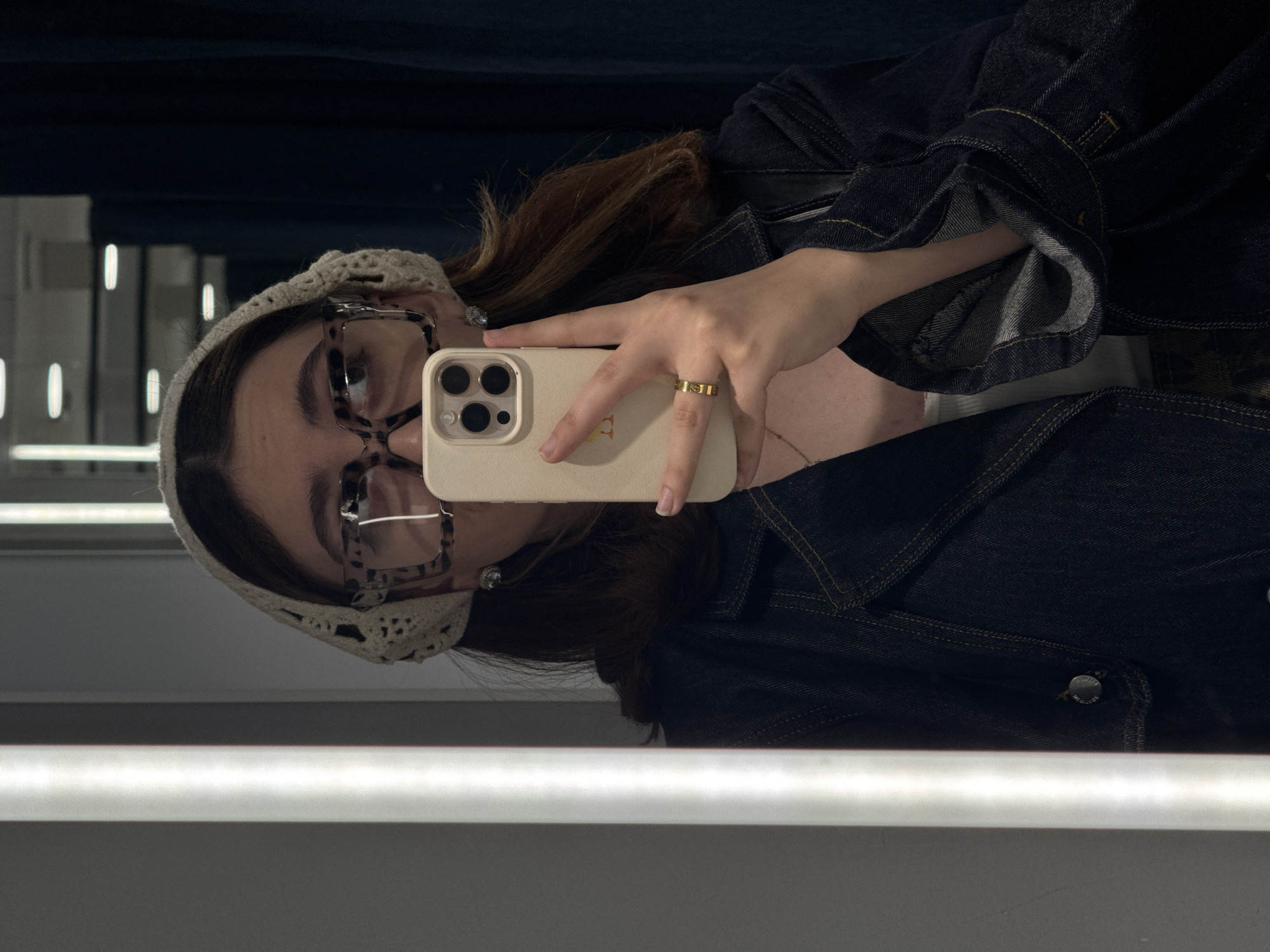The image shows a person taking a selfie in a mirrored environment. The individual has brown hair and is wearing glasses and a crocheted headband. They are holding a beige smartphone. They also appear to be wearing a dark denim jacket. The setting seems to be a dressing room or similar retail space, evidenced by the reflective surface and vertical lighting fixtures in the background. There's no information in the image to suggest it was taken in Yerevan, Armenia.