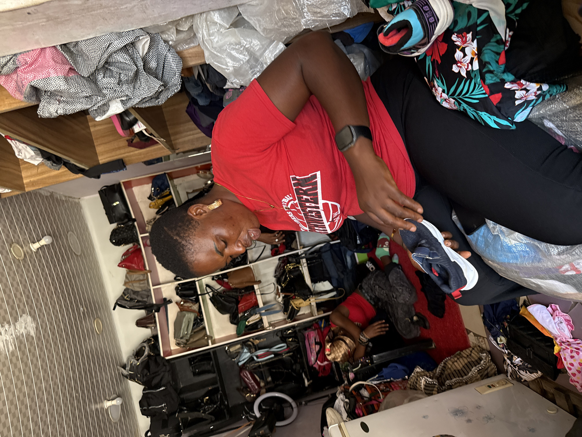 This is a medium shot of a crowded shop in Abuja, Nigeria, with a woman in a red t-shirt and black leggings in the foreground. She is holding a blue and white sneaker, examining it. The shop is filled with shelves packed with handbags and clothing, and there is another person lying on the floor in the background, wearing a red tank top and gray pants. The shop appears to be dimly lit, with fluorescent lights on the ceiling. The overall impression is one of a busy, possibly informal retail environment.