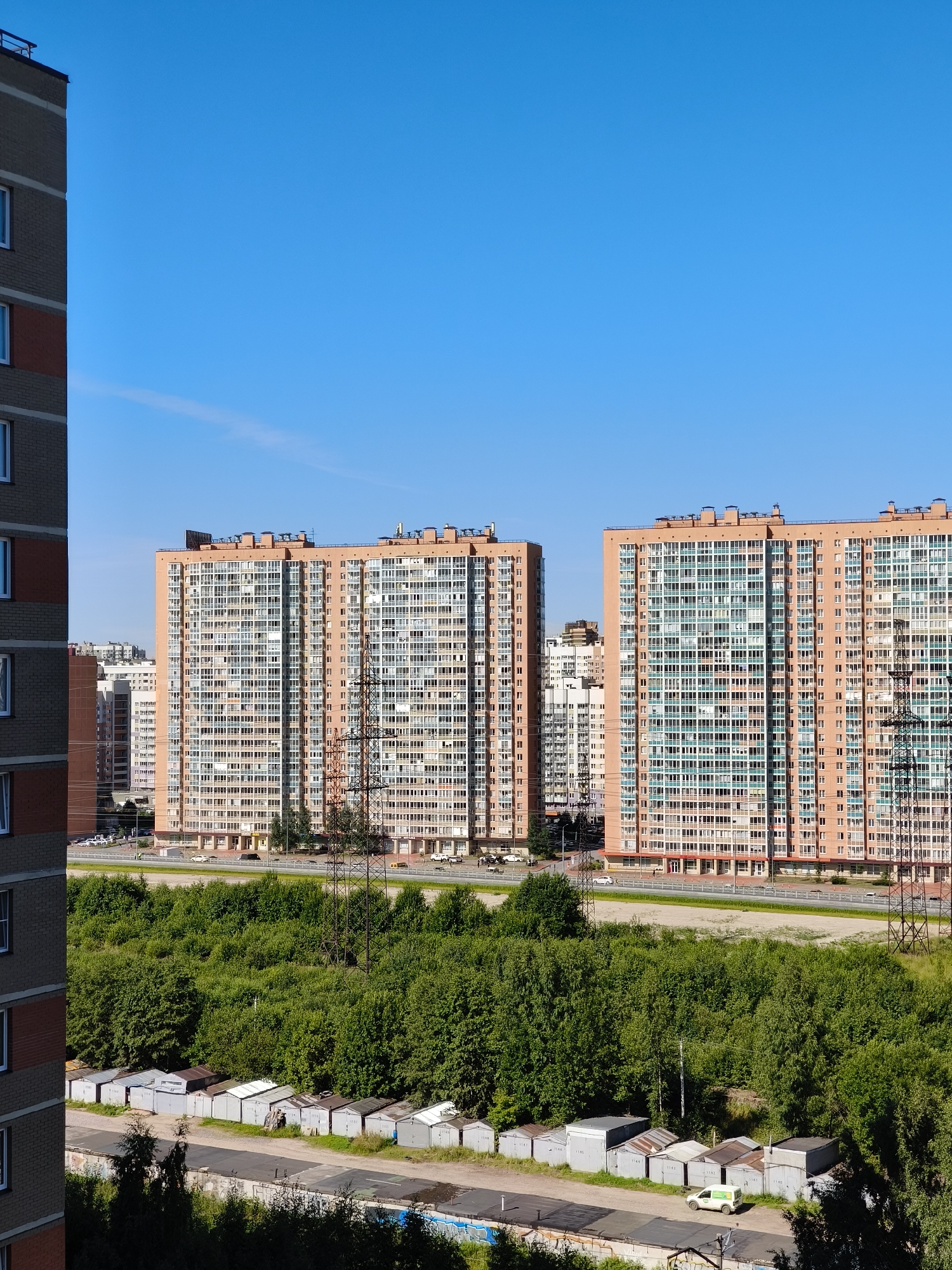 The image captures a view of Murino, Russia, on a clear day. In the foreground, a tall, modern brick building with light-colored window frames is partially visible on the left. Beyond this, two large, light-orange apartment buildings with numerous windows dominate the mid-ground. These buildings appear to be part of a new residential complex. Between the apartment buildings and the foreground structure, a wide, paved road with traffic is visible. A dense line of green trees and foliage separates the road from a row of small, uniform garages situated in the lower part of the image. One white car with green accents is parked near the garages. In the background, the sky is a clear, bright blue. Power lines and transmission towers are also present, indicating urban infrastructure.
